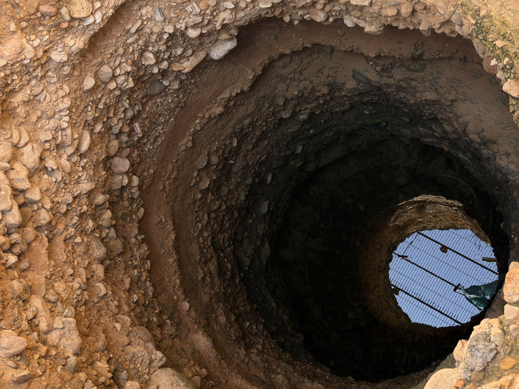 View looking down a well in Sant Martí Sarroca, Spain. The well is constructed of earth and stone, with a metal grate covering the opening at the bottom, through which blue sky is visible.