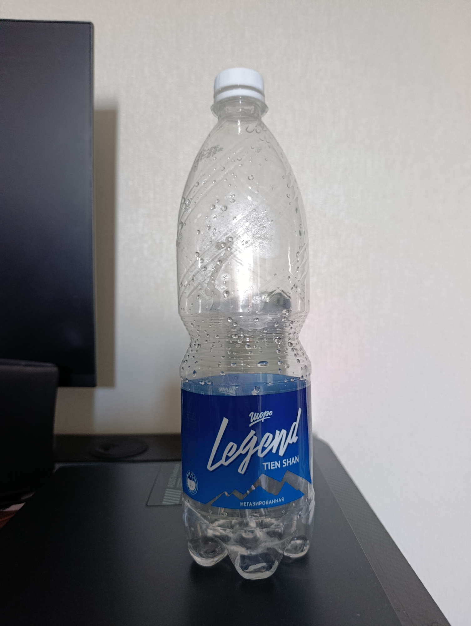 An empty plastic water bottle with a white cap sits on a black surface. The bottle is mostly clear with condensation on the outside. A blue label with the word "Legend" in white script and "TIEN SHAN" in smaller white text is visible on the lower portion of the bottle. Below "TIEN SHAN" is the word "НЕГАЗИРОВАННАЯ" in white text. The background is a plain, lightly textured off-white wall. To the left of the bottle is the side of a dark, rectangular object, possibly a monitor or a cabinet. The scene is brightly lit, casting a subtle shadow from the bottle onto the surface it rests on.