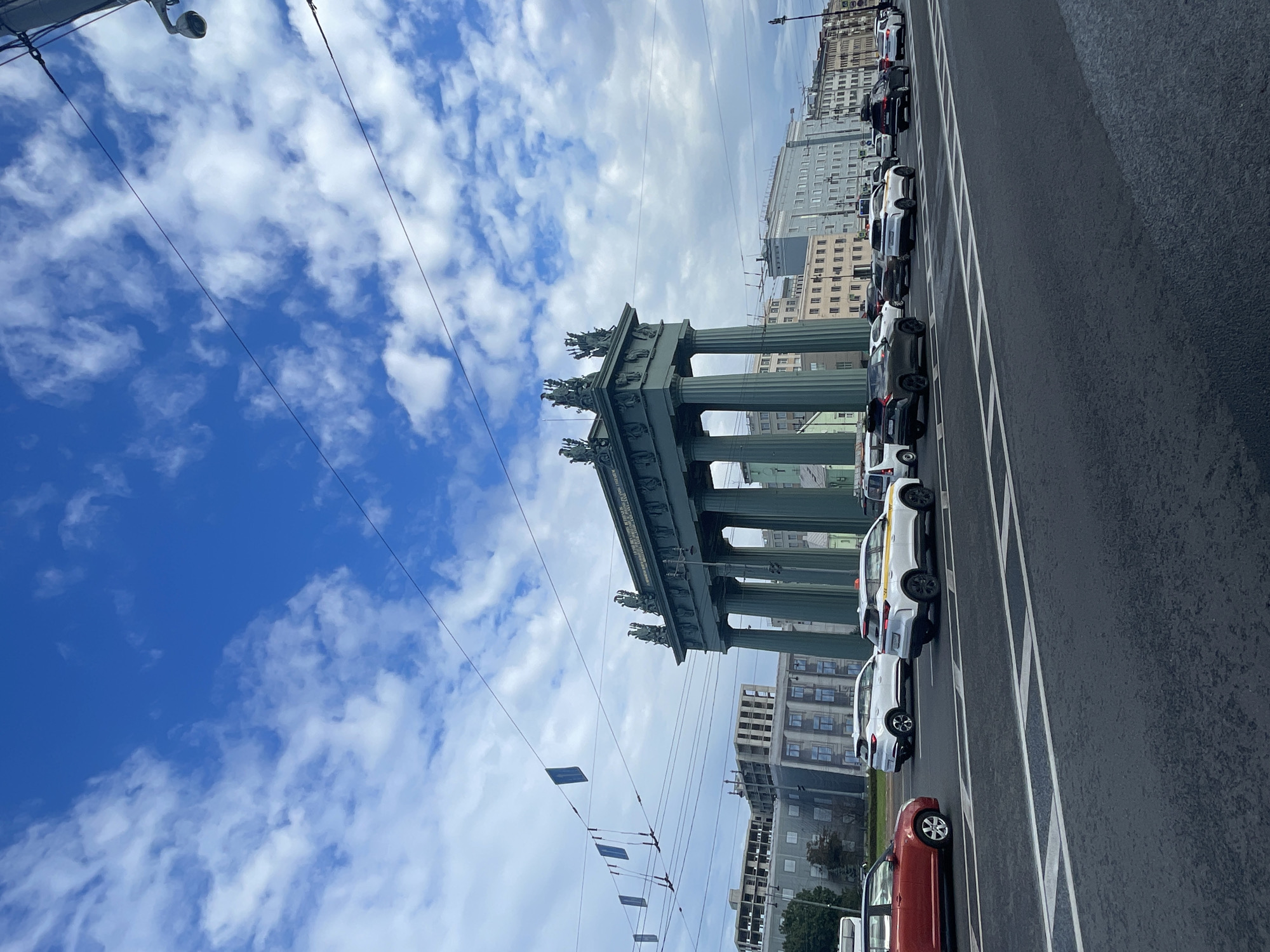 The image depicts the Rostral Columns in Saint Petersburg, Russia, on a cloudy day with a bright blue sky visible. The columns, a prominent architectural landmark, are in the background, their tall, fluted structures supporting the upper architectural elements. In the foreground, a multi-lane asphalt road is filled with vehicles, including several cars and what appears to be a taxi. The cars are in motion, suggesting traffic. Buildings line the street behind the columns, showcasing a mix of architectural styles. Overhead wires and a security camera are also visible, indicating an urban environment. The overall scene captures a slice of daily life in the city, with historical architecture set against a backdrop of modern transportation and infrastructure.