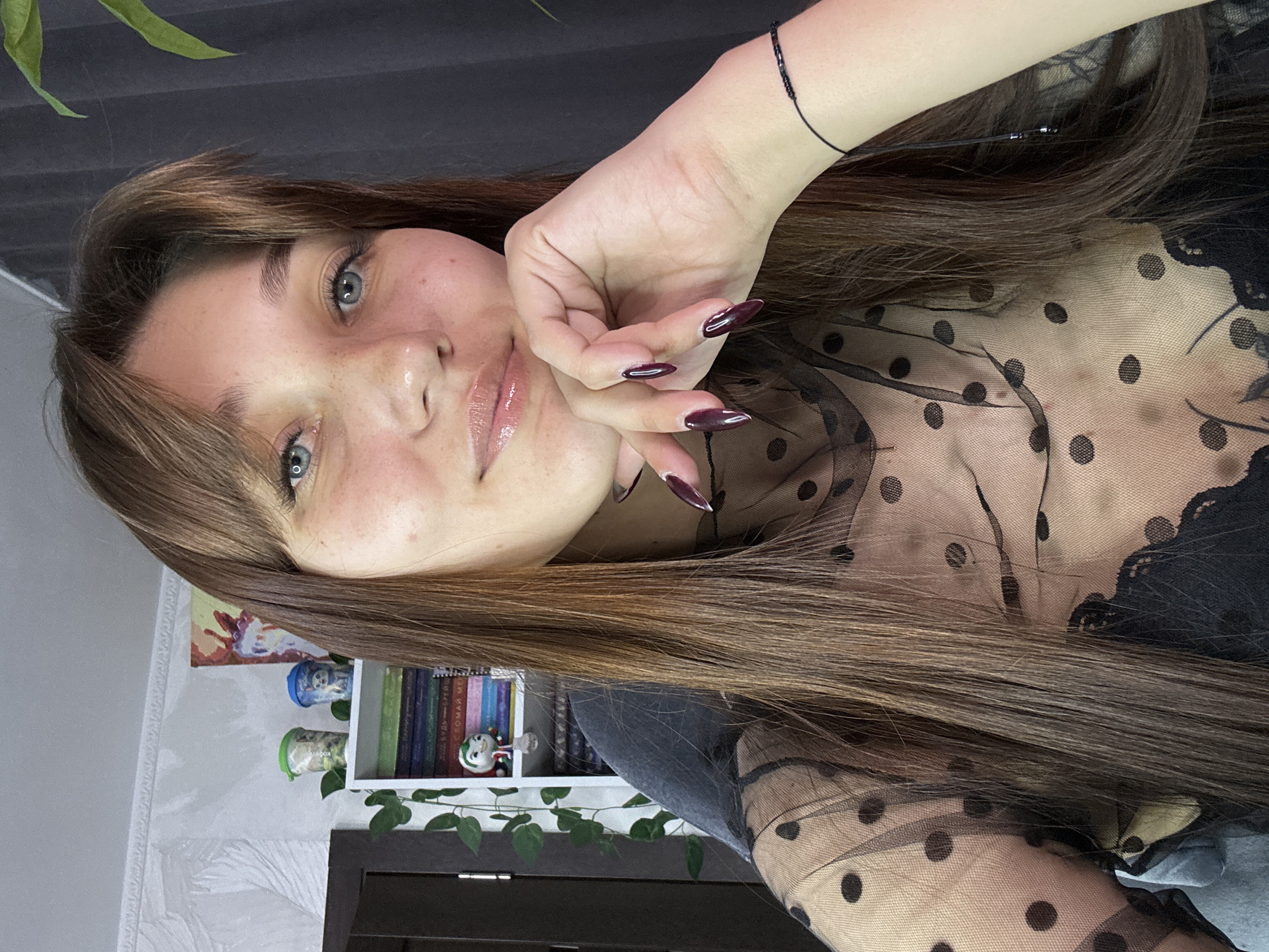 A young woman with long, dark brown hair and light blue eyes is pictured in a selfie. She has a light dusting of freckles across her nose and cheeks and her lips are glossy. She is resting her chin on her right hand, which has dark purple, pointed fingernails. She is wearing a sheer black polka dot top over a black lace bra. In the background, a white bookshelf is visible, adorned with small figurines and what appear to be books. A plant with green leaves is also present in the background, partially obscuring a dark gray curtain. The wall behind the bookshelf has a textured pattern. The setting appears to be an indoor room, potentially a home office or living space, in Novokuznetsk, Russia.
