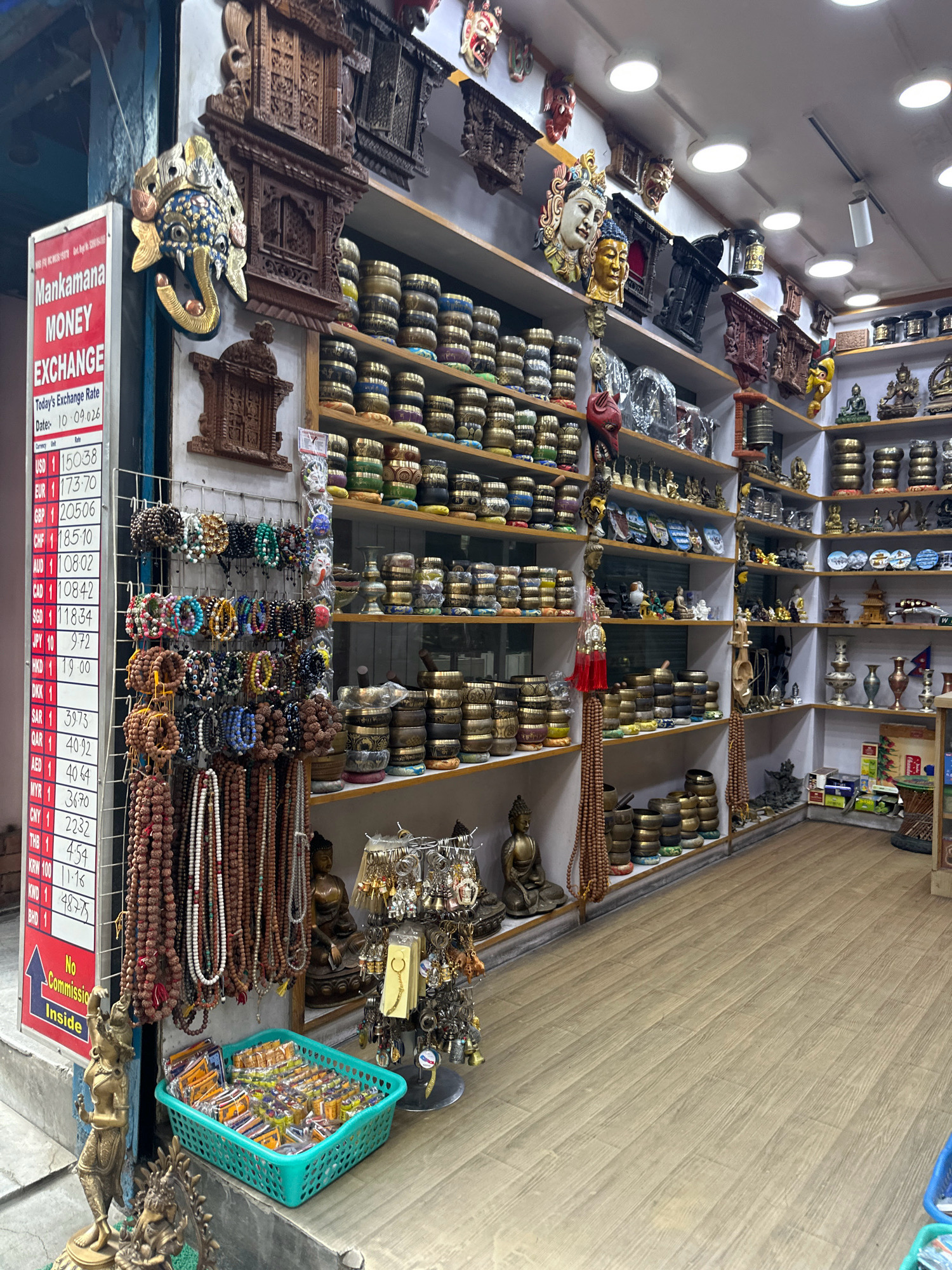This is a wide-angle, eye-level, interior shot of a shop in Kathmandu Metropolitan City, Nepal, selling religious and decorative items. The shop is filled with shelves displaying numerous products, including prayer bowls, statues, masks, and bracelets.
In the foreground, on the left, is a money exchange board with currency rates. Below the board, a turquoise basket is filled with small, individually packaged items. To the left of the basket, a brass statue of a standing woman is visible.
The shop's walls are lined with shelves. The upper shelves hold carved wooden items and decorative masks, including a prominent blue elephant mask with gold accents. The middle shelves are filled with stacks of prayer bowls, while lower shelves display more prayer bowls, statues of Buddha, and various religious artifacts. A rack of hanging bracelets is also visible on the left side of the shop.
The floor is made of light brown laminate wood planks. The lighting is provided by recessed ceiling lights, some of which are visible in the upper part of the image. The overall impression is of a crowded but organized retail space dedicated to cultural and spiritual merchandise.