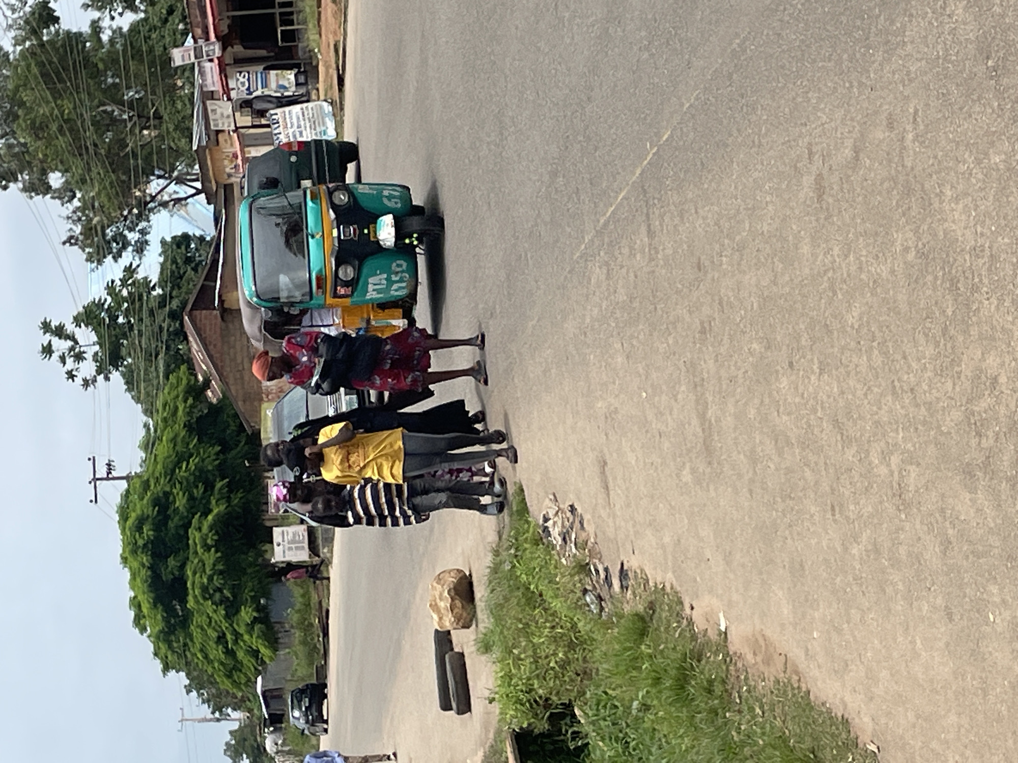 The image depicts a street scene in Jos, Nigeria, under an overcast sky. In the foreground, a dirt and asphalt path runs along the side of a paved road. Lush green grass and some debris line the edge of the path. Two discarded tires and a large rock are visible on the side of the road.
On the road, a group of people are walking. A young person in a striped shirt and grey pants is at the front, followed by a person in a yellow t-shirt and grey pants. Behind them, several other individuals are walking, some obscured by others. One woman wears a red and white patterned dress and an orange head covering, carrying a dark bag. Another woman is dressed in a black outfit with a patterned headscarf.
Further down the road, a teal and yellow auto-rickshaw, also known as a "Keke Napep," is the most prominent vehicle. It has markings that include "PTA" and "1150" on the front. A dark green SUV is behind the auto-rickshaw.
In the background, buildings with corrugated metal roofs and some shops line the street. A tall wooden utility pole with wires is also visible. The overall environment suggests a typical roadside setting in a Nigerian city.