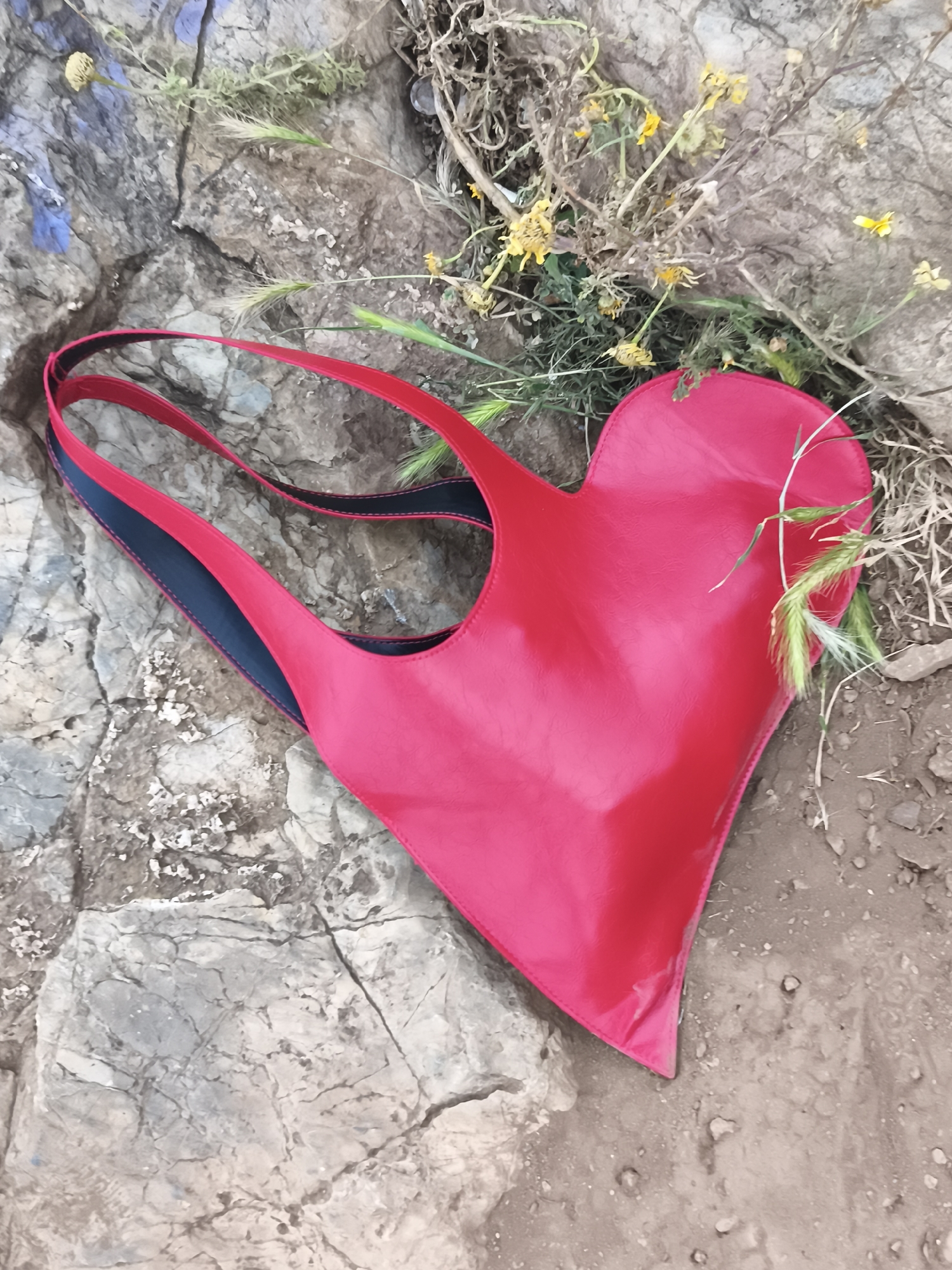 The image shows a heart-shaped red leather bag with black lining in the straps, lying on a surface that appears to be a combination of rocky terrain and dirt. Some dried yellow flowers and green blades of grass are near the bag. The rocky surface has a grayish-white hue with cracks and textures. The dirt portion of the surface is a light brown color. The location is unspecified but could plausibly be in Barcelona, Spain, based on the Mediterranean vegetation and stone materials present, but this is just speculative.
