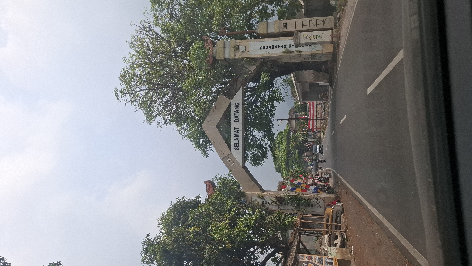The image shows an overhead archway spanning a road in Sukasari, Indonesia, with the text "SELAMAT DATANG" written on it, meaning "Welcome." The archway is made of concrete and decorated with traditional Indonesian motifs. On the left side of the road, there is a small structure with some items displayed outside, possibly a shop or stall. Several vehicles, including motorcycles and a car, are seen on the road, with some people riding them. The road is paved and has white markings. On the right side of the archway, there is another concrete pillar with text and graffiti on it. The environment appears to be semi-urban with trees and buildings visible in the background. The sky is overcast.