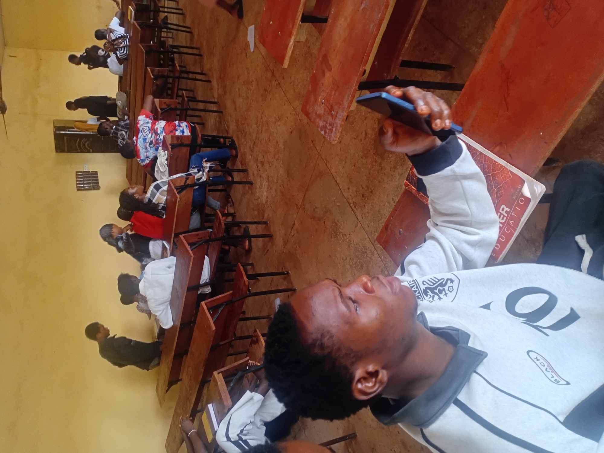 A classroom in Unknown, Unknown is shown, filled with students sitting at wooden desks. A person in the foreground is looking at a cell phone in their hand. The other students appear to be engaged in some sort of classroom activity. The classroom walls are a pale yellow.