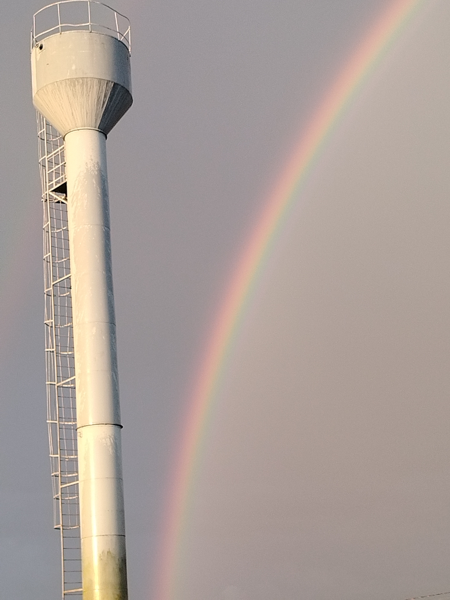 An outdoor, medium shot captures a tall, white water tower with a ladder on its left side, positioned against a soft gray sky. To the right of the tower, a faint rainbow arcs across the sky. The water tower is cylindrical with a conical top and a railing around its uppermost section. The ladder is attached vertically to the side of the tower, extending from near the bottom to the top. The sky is overcast, creating a diffused light. The scene appears to be located in the Завуццеўскі сельскі Савет region of Belarus.