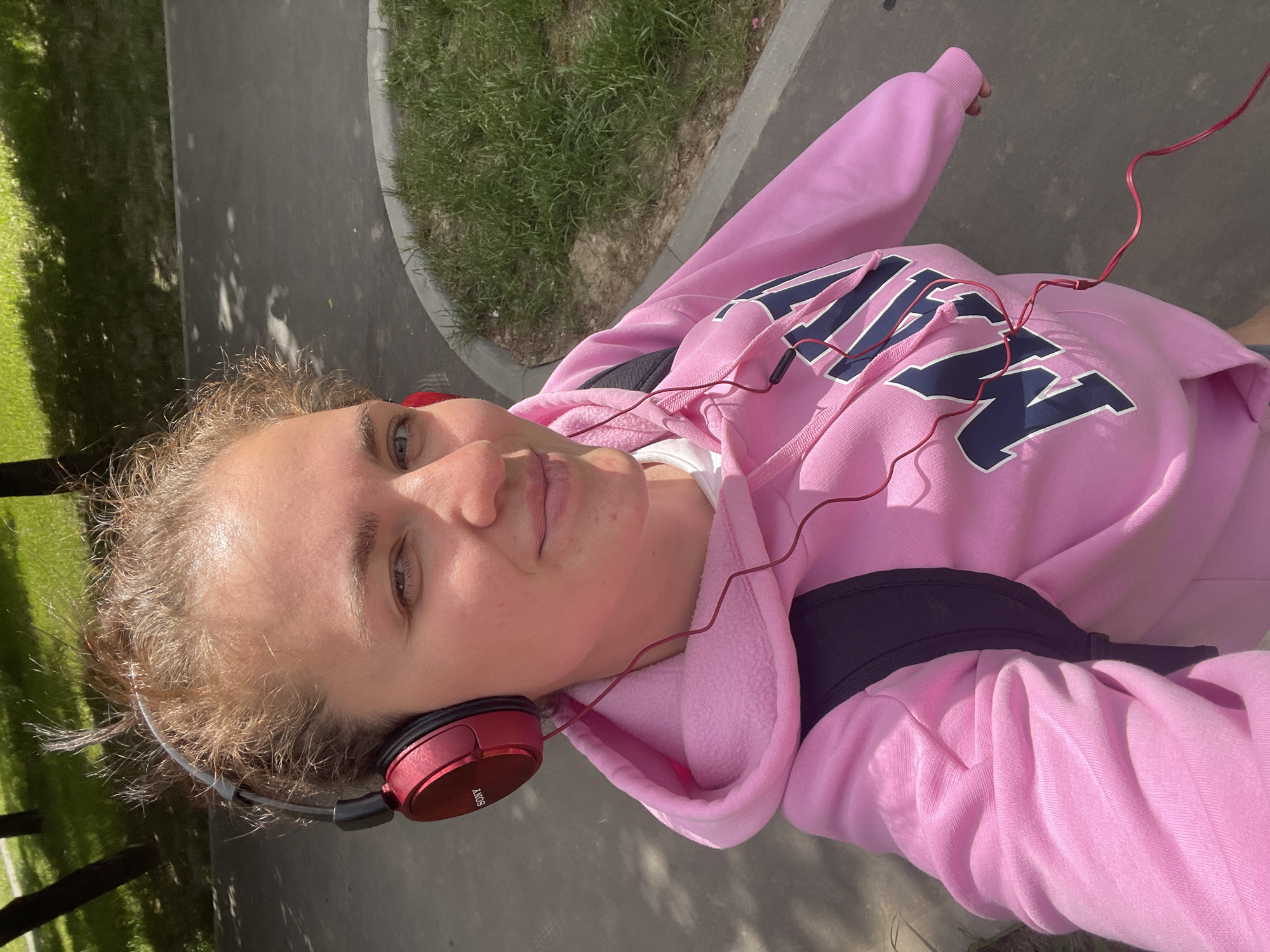A young person wearing a pink hoodie and red headphones is taking a selfie outside on a sunny day. They are standing on a gray paved surface next to a patch of green grass. The location is Moscow, Russia. The person is wearing a black backpack.