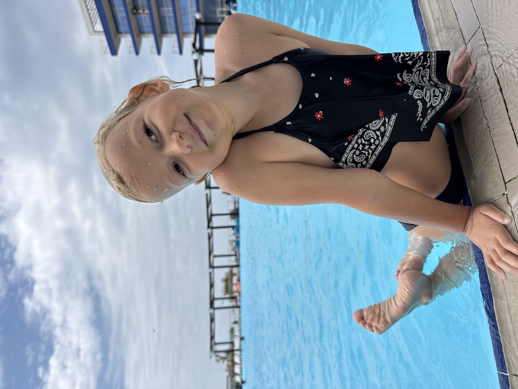 A young girl with wet blonde hair and a black swimsuit with a paisley and floral pattern smiles as she rests on the edge of a swimming pool. Her left arm is extended along the pool's tiled edge, and her left hand is visible. Her right arm is bent, with her hand near her head. One of her feet is submerged in the bright blue water. The background shows a cloudy sky and a blurred structure that appears to be a building with a modern, multi-level design. The scene suggests a warm day at an outdoor swimming pool.