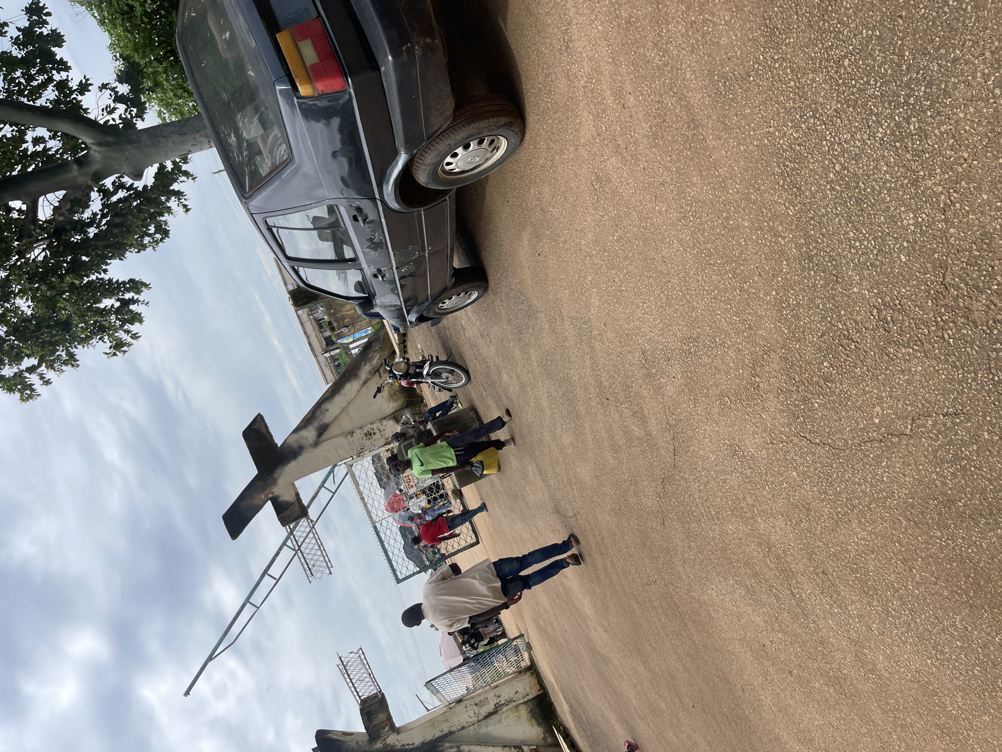 In Rafin Par, Nigeria, a black Volkswagen hatchback is parked on a paved lot. The car is facing right, with its rear driver's side tire visible. The car's paint is worn and scratched, and the taillight assembly is red and yellow. In the background, several people are walking near an entrance structure featuring a cross. One person, wearing a light-colored shirt and blue jeans, walks away from the camera towards the entrance. Another person in a green shirt carries a yellow bag. A motorcycle is parked to the right of the entrance. The sky is cloudy.