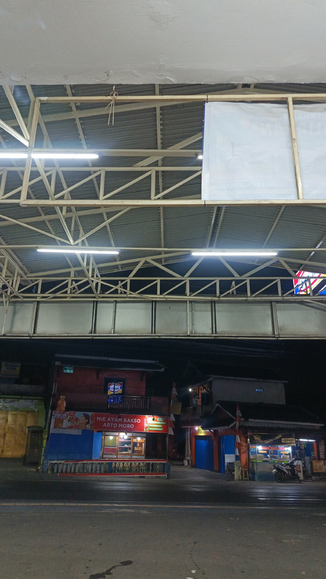 The image depicts an exterior view of a street scene at night, likely in Margajaya, Indonesia. The upper portion of the image shows the underside of a covered walkway or structure with a corrugated metal roof supported by a metal truss system. Fluorescent lights are visible, illuminating the area. A large white banner is attached to the structure on the right side, displaying some text, though it is partially obscured and not clearly legible.
Below the walkway, a row of small shops or food stalls is visible. The most prominent stall in the center is red and blue, with signage that reads "MIE AYAM BAKSO ARTO MORO". This stall appears to be open for business, with lights on inside and visible seating and food preparation areas. To the left and right of this stall are other similar structures, some with blue doors and illuminated interiors. A motorcycle is parked in front of one of the stalls on the right. The street in front of the shops is wet, suggesting recent rain. The overall atmosphere is that of a commercial area at night. There are no people clearly visible in the image.