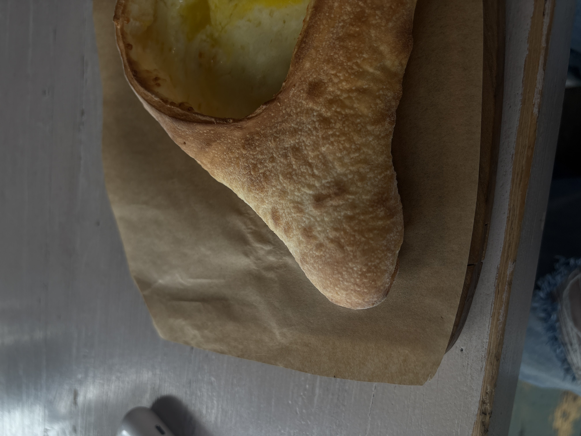 A close-up, overhead shot shows a khachapuri, a Georgian cheese-filled bread, resting on brown parchment paper on a light-colored wooden table. The khachapuri is boat-shaped, with a golden-brown crust and a molten, yellow cheese filling. The table is slightly worn and shows texture. In the upper left corner, a white wireless earbud case is partially visible. The background is blurred, but suggests an indoor setting. The bottom of the image shows a hint of a person's legs, wearing distressed jeans. The scene is devoid of specific context related to Krasnogorsk, Russia.