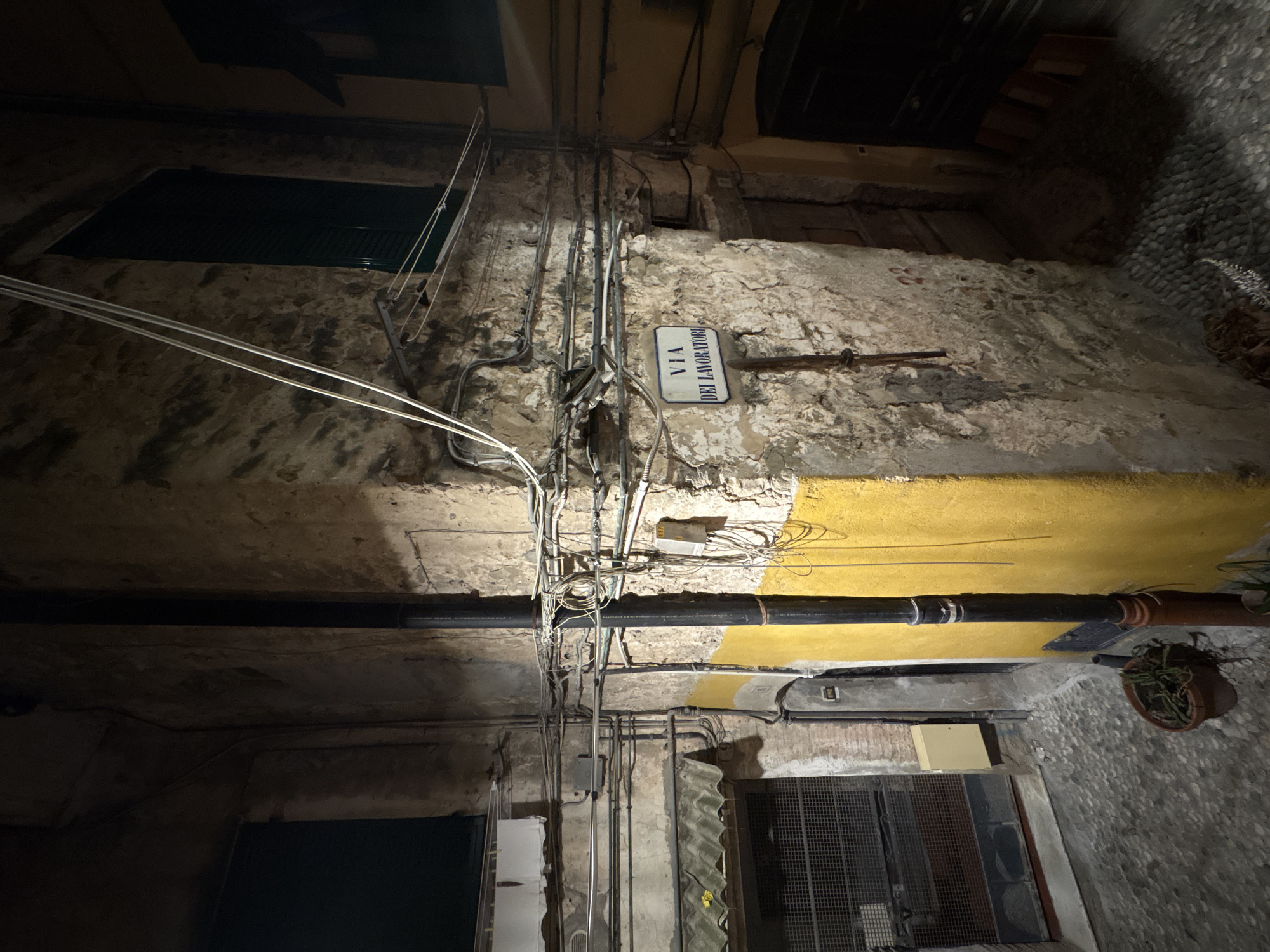 The image shows a narrow alleyway in Sanremo, Italy, at night. The walls of the buildings are made of stone and plaster, with visible signs of wear and tear. A street sign reading "VIA DEI LAVORATORI" is mounted on the wall. Various electrical wires and conduits run along the buildings. In the foreground, there is a cobblestone street and a potted plant. A dark-colored pole, possibly a lamppost or drainpipe, runs vertically along the wall. The lighting suggests it is nighttime, with artificial light sources illuminating parts of the alley.