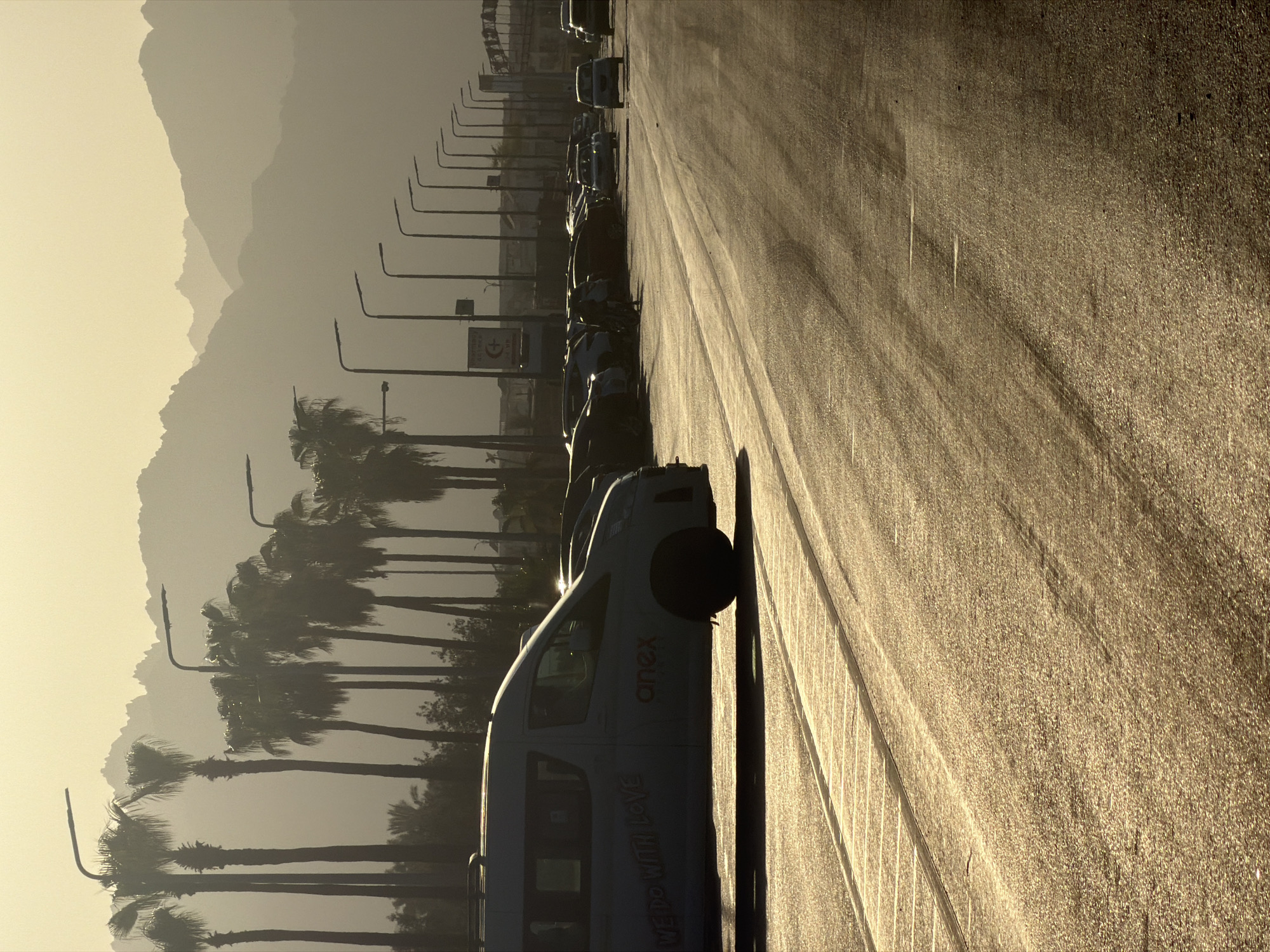 In Sharm El Sheikh, Egypt, at a parking lot bathed in the golden light of sunset, a row of palm trees stands silhouetted against a hazy sky. In the foreground, a white van with "WE DO WITH LOVE" and "onex" written on its side is parked. Beyond the van, a line of cars is visible, and in the distance, the undulating shapes of mountains form a majestic backdrop. Streetlights line the edge of the parking lot, their designs simple and modern. The overall atmosphere is one of tranquility and warmth, as the day draws to a close in this popular Egyptian tourist destination.