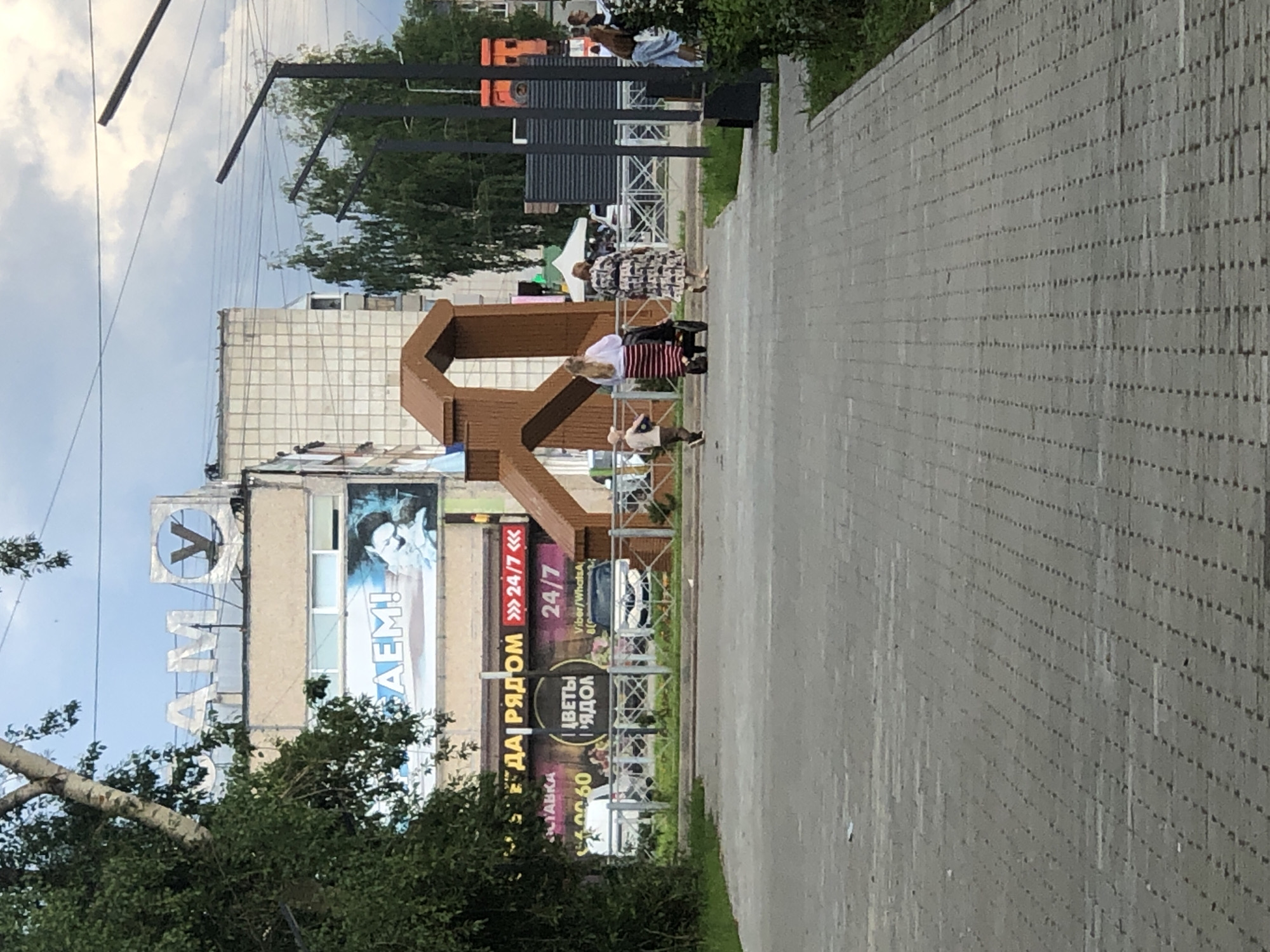 The image shows a street scene in Solikamsk, Russia, featuring a large building with signage. The building's facade has large letters spelling "САМ У" at the top, and a banner with the text "АЕМ!" below. Further down, a sign displays "ЕДА РЯДОМ" (Food Nearby) with "24/7" in red numerals, and another sign shows "ЦВЕТЫ РЯДОМ" (Flowers Nearby) also with "24/7". In the foreground, a paved walkway leads towards the building. A wooden, abstract sculpture is positioned in front of the building. Several people are visible: a child running, a woman with a stroller, and other individuals walking in the distance. The environment appears to be an urban setting with trees and streetlights.