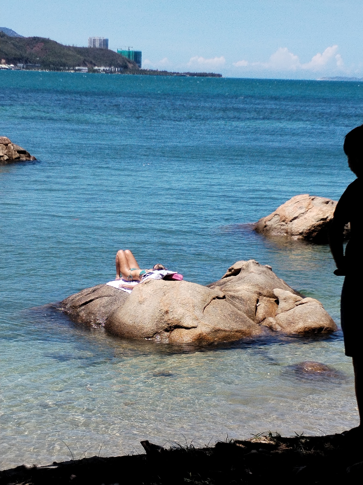 The image depicts a tropical scene at a beach in Bac Nha Trang Ward, Vietnam. A person lies prone on a large rock in the water, appearing to be sunbathing. They are wearing a bikini and resting on a white towel. A second person, silhouetted, stands at the edge of the beach to the right. Clear water surrounds the rocks, and in the distance, a coastline with buildings and hills is visible under a clear sky.