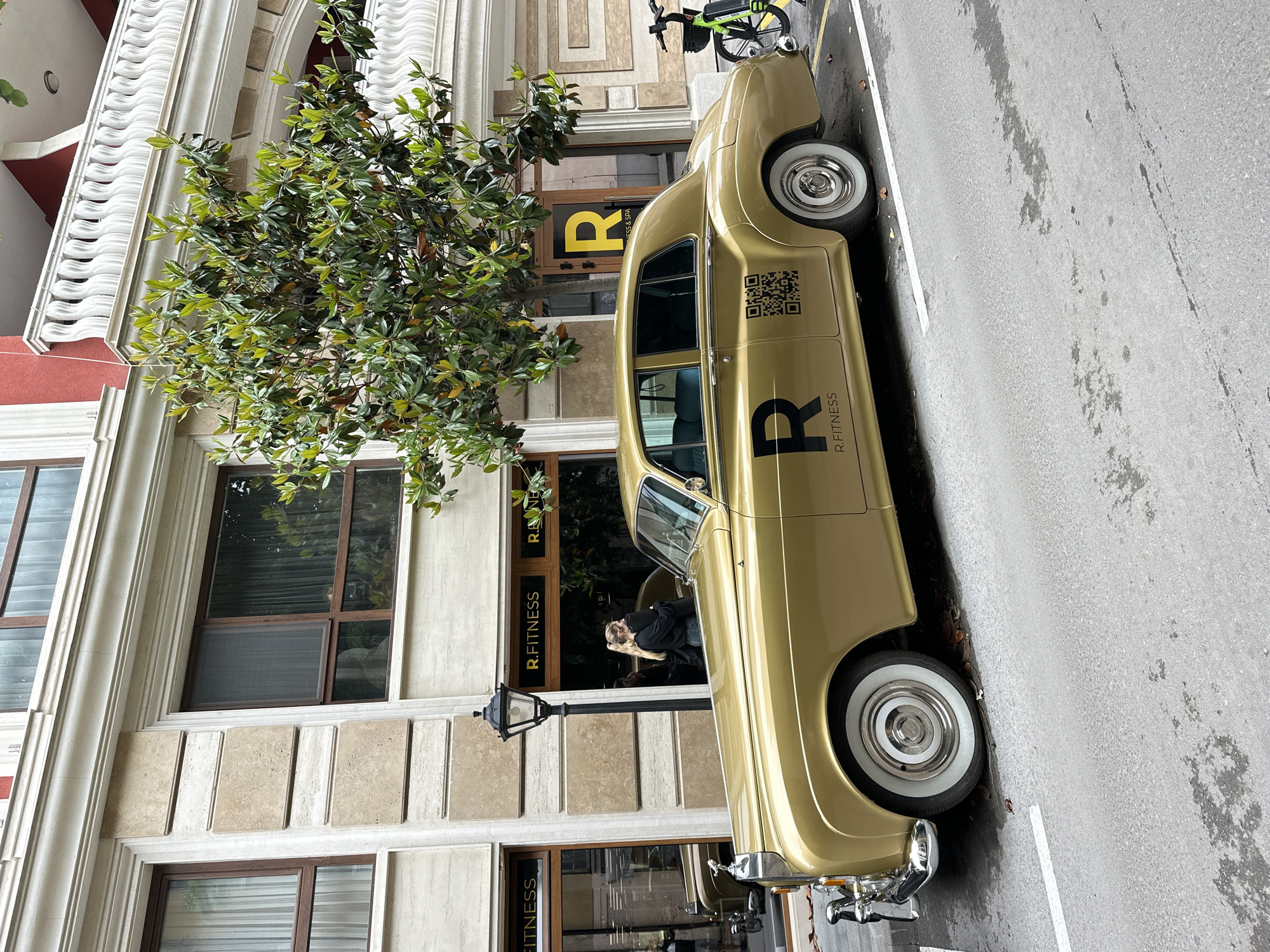 An eye-level, full shot captures a gold-colored vintage sedan, possibly a GAZ-21 Volga, parked alongside a stately building on an urban street in Varna, Bulgaria. The car, adorned with prominent chrome bumpers and whitewall tires, features large black text on its side reading "R. FITNESS" and a large stylized "R." Above this text, a black QR code is also visible.
A blonde woman, dressed in dark clothing, leans out of the front passenger window of the car, looking towards the left side of the frame with a relaxed posture. The car is parked on a paved street, which shows some damp patches, suggesting recent moisture or an overcast day. A white parking line is visible on the asphalt near the car's front tire.
The building behind the car exhibits classical architectural elements, including light-colored stone or stucco with decorative moldings, arched windows, and a prominent entrance. The entrance features a large glass door and window with "R. FITNESS" displayed in black text, mirroring the car's branding. Above this, "FITNESS & SPA" is also visible. A lush green tree with dense foliage partially obscures the building's facade and the car. A black ornate street lamp is positioned in front of the building. In the upper background, a bright green bicycle, possibly a shared or rental bike, is parked on the sidewalk. The scene appears to be set during daytime, under what seems to be diffused or overcast natural light.
