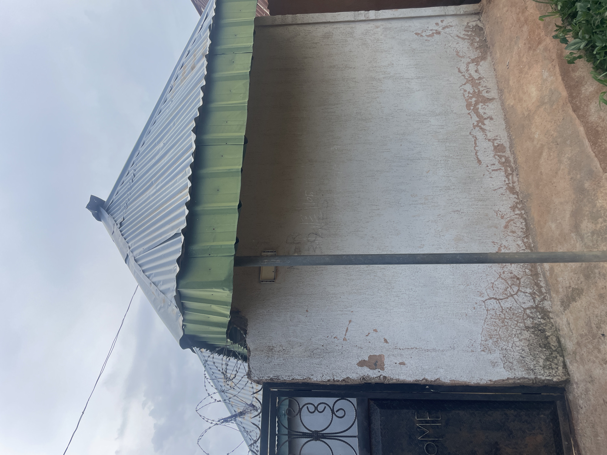 The image depicts the exterior of a building in Bukuru, Nigeria. The building features a corrugated metal roof, partially green and partially silver, with a visible seam at the peak. Barbed wire is stretched across the top of the wall on the left side of the image, over a black metal gate with ornate scrollwork. The word "OME" is visible on the gate. The wall itself is textured and appears to be painted white, with areas of peeling plaster revealing an underlying reddish-brown material. There are some faint markings or graffiti on the wall. A vertical gray pole is positioned in front of the wall. The ground in front of the building is a mix of dirt and concrete. Green vegetation is visible in the lower right corner. The sky is overcast with clouds.