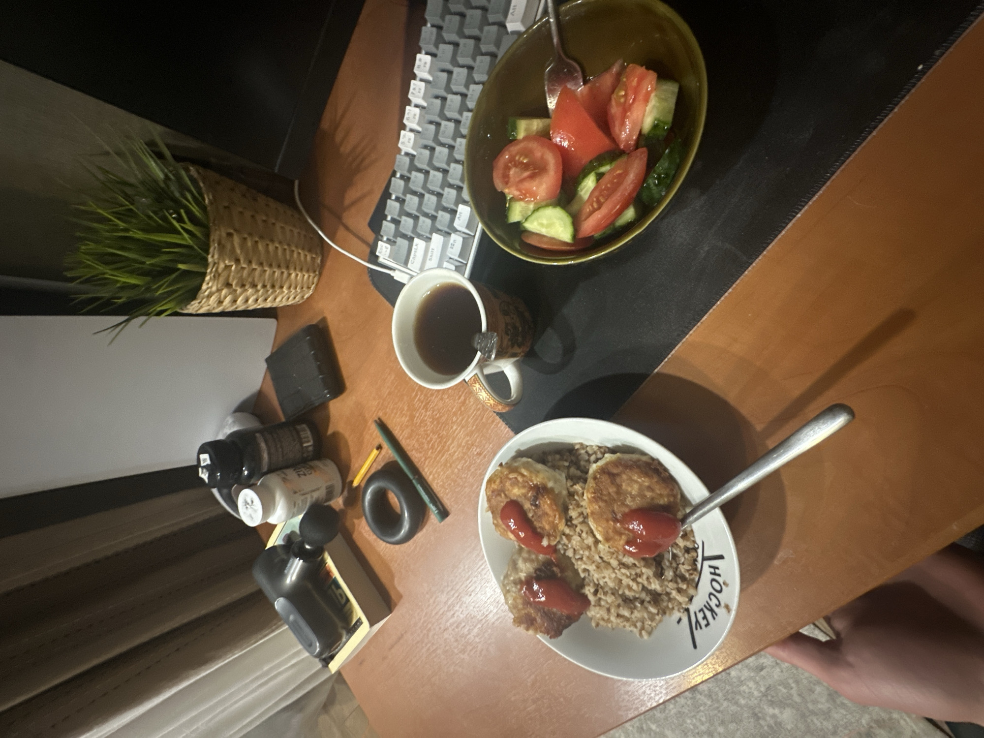 The image shows a desktop with various items arranged on it. A keyboard and a mousepad with a bowl of salad are positioned on the upper right. A mug of coffee is located below the bowl. Below the mug and to the left are several small bottles, a black ring, and a pen. In the lower center is a white plate filled with food, with the word "HOCKEY" visible on the plate's rim. A silver spoon is also on the plate. The location of the image is Unknown, Unknown.