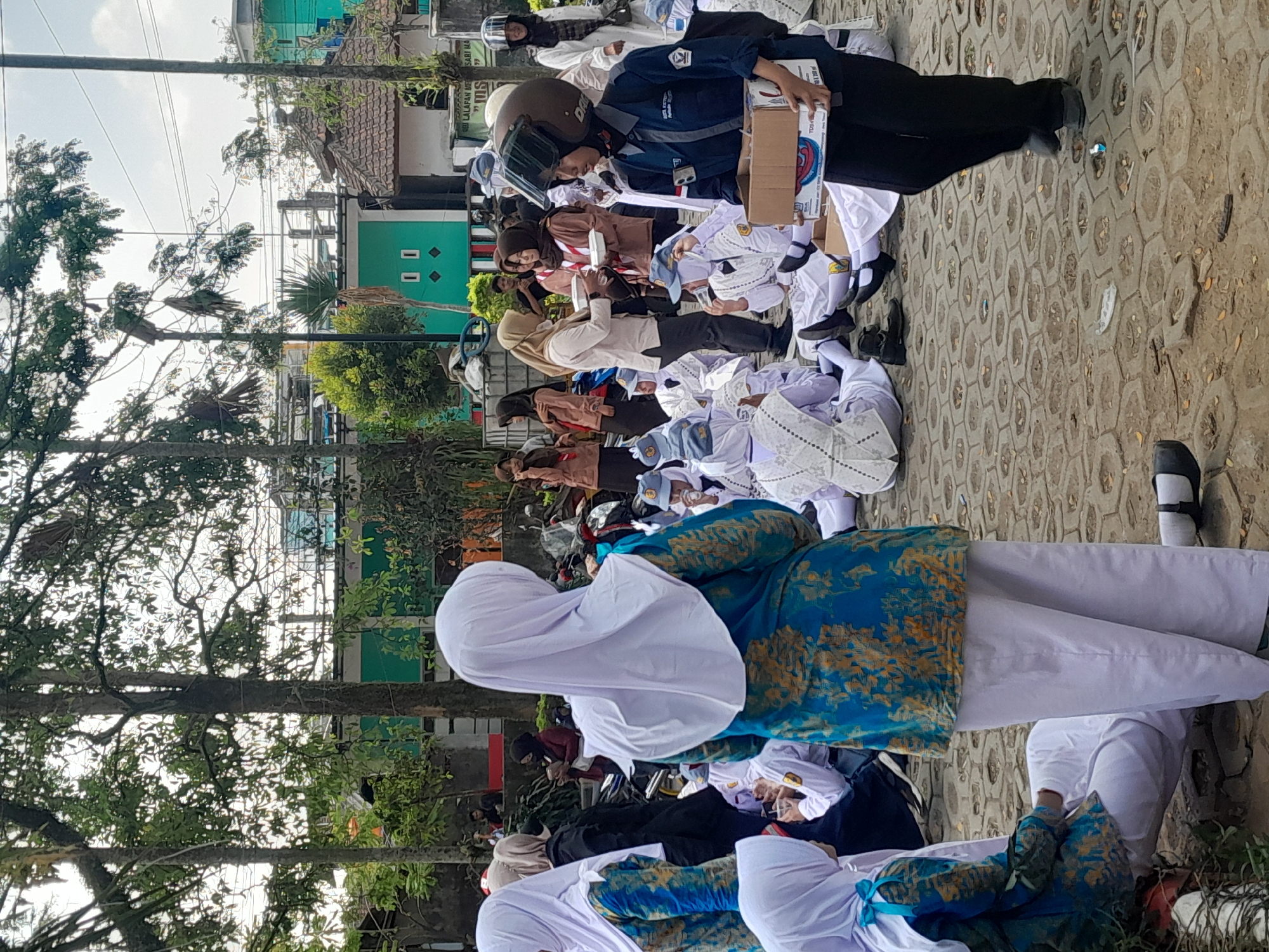 In Genteng Kulon, Indonesia, a group of students, many wearing white uniforms and headscarves, are gathered outdoors on a paved area with a hexagonal pattern. Some students are sitting on the ground, while others are standing. A young woman wearing a dark blue uniform, a helmet, and a mask is walking towards the right, holding a cardboard box. She appears to be distributing or collecting items. Other individuals in the background are also visible, some in uniforms and others in casual clothing. The environment includes trees, buildings with colorful facades, and power lines, suggesting an urban or semi-urban setting. The atmosphere appears to be a casual gathering or event.