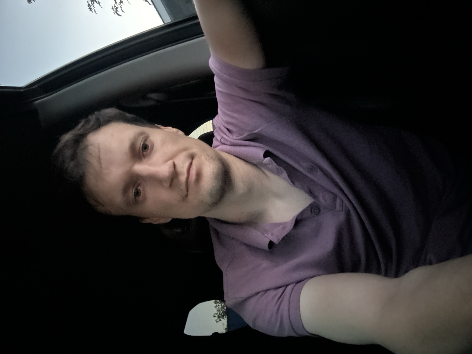 A man is sitting in the driver's seat of a car. He is wearing a purple polo shirt. The car interior is dark. Through the driver's side window, a light-colored sky and some trees are visible. The scene appears to be outdoors, possibly in Shatava, Ukraine, during dusk or dawn. The man is looking towards the camera, and his right arm is extended towards the passenger side. His expression is neutral.