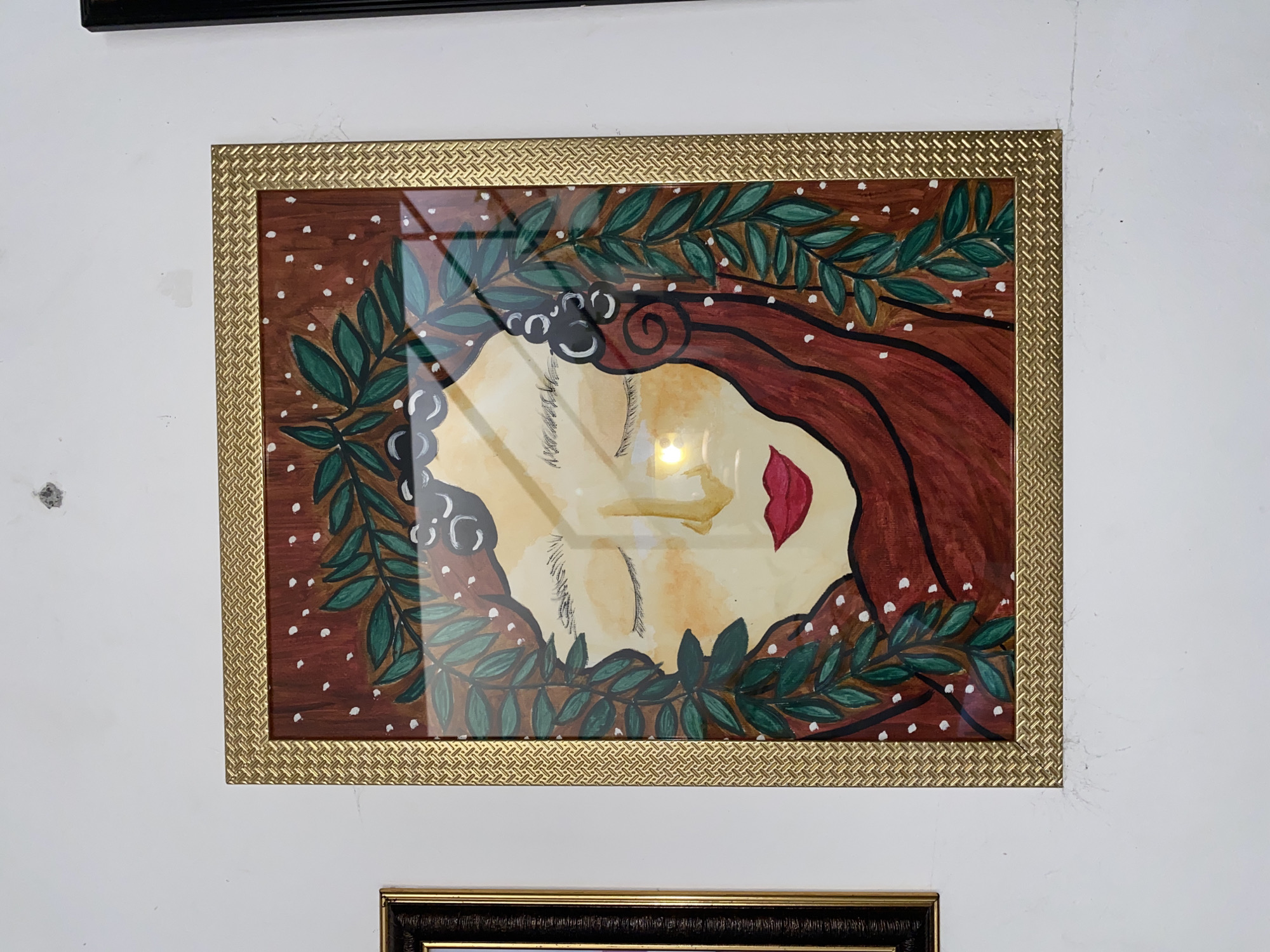 The image shows a framed painting of a woman's face with closed eyes, adorned with green leaves and white dots, hanging on a white wall. The painting is in a gold frame with a textured pattern. To the left of the framed painting is another framed artwork with a black frame. The wall appears to be slightly textured and has some minor marks. The scene is located in Davao City, Philippines.