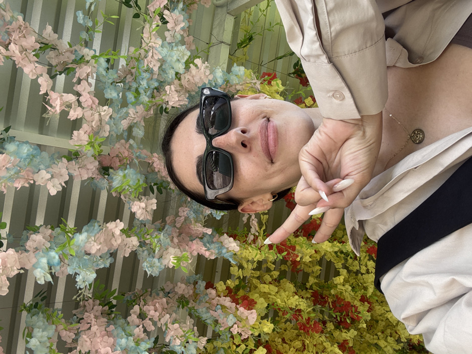 A person with dark hair pulled back, wearing dark sunglasses and a light-colored shirt, poses against a backdrop of flowers. The background features pastel blue and pink blossoms mixed with yellow and red flowers. The person makes a peace sign with their hand, displaying long, light-colored nails. A gold necklace with a pendant is visible. The photograph's perspective appears to be taken from below and slightly angled.
