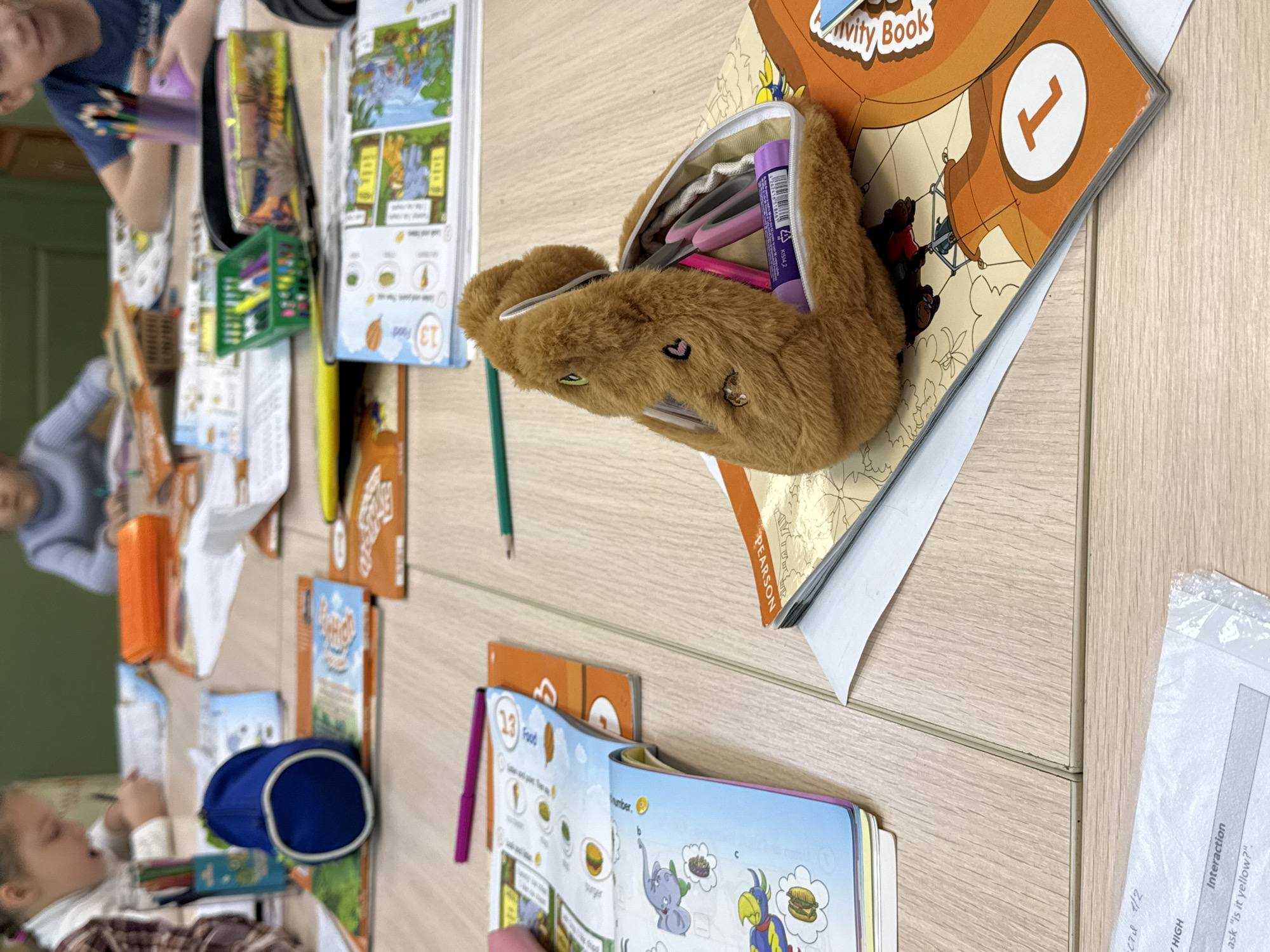 The image depicts a classroom setting, likely in the city of Perm, Russia, given the Cyrillic text on some of the materials. Several children are seated around a light-colored wooden table, engaged in activities with books and writing implements. A fuzzy, brown pencil case overflowing with writing tools rests on top of an open book titled "Activity Book." A textbook with images of food and numbers is also visible. Other books, pencil cases, and baskets filled with crayons are scattered across the table, suggesting a group learning environment. Children are partially visible at the table, indicating an interactive classroom scenario. A printed worksheet is located to the far right of the image.