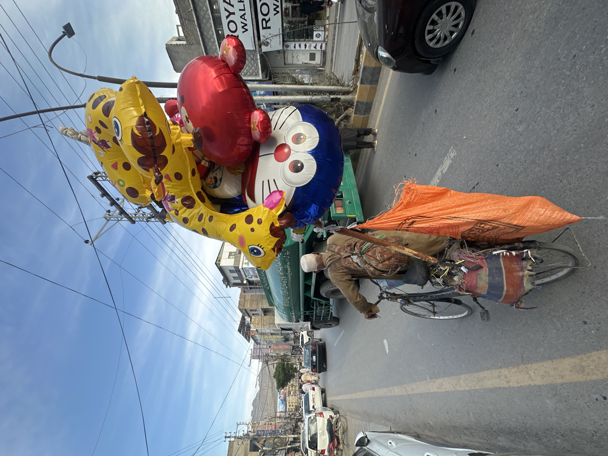 A vendor on a bicycle laden with goods and a large assortment of balloons is riding down a street in Quetta City Tehsil, Pakistan. The balloons are cartoon character shaped, with yellow cheetah-like balloons, a red balloon, and a blue Doraemon character balloon. The bicycle also carries an orange sack. The man is wearing a hat and jacket.
The street is lined with parked cars, moving vehicles, and buildings. Signage and storefronts indicate commercial activity. Power lines crisscross the sky. In the background, mountains are visible.