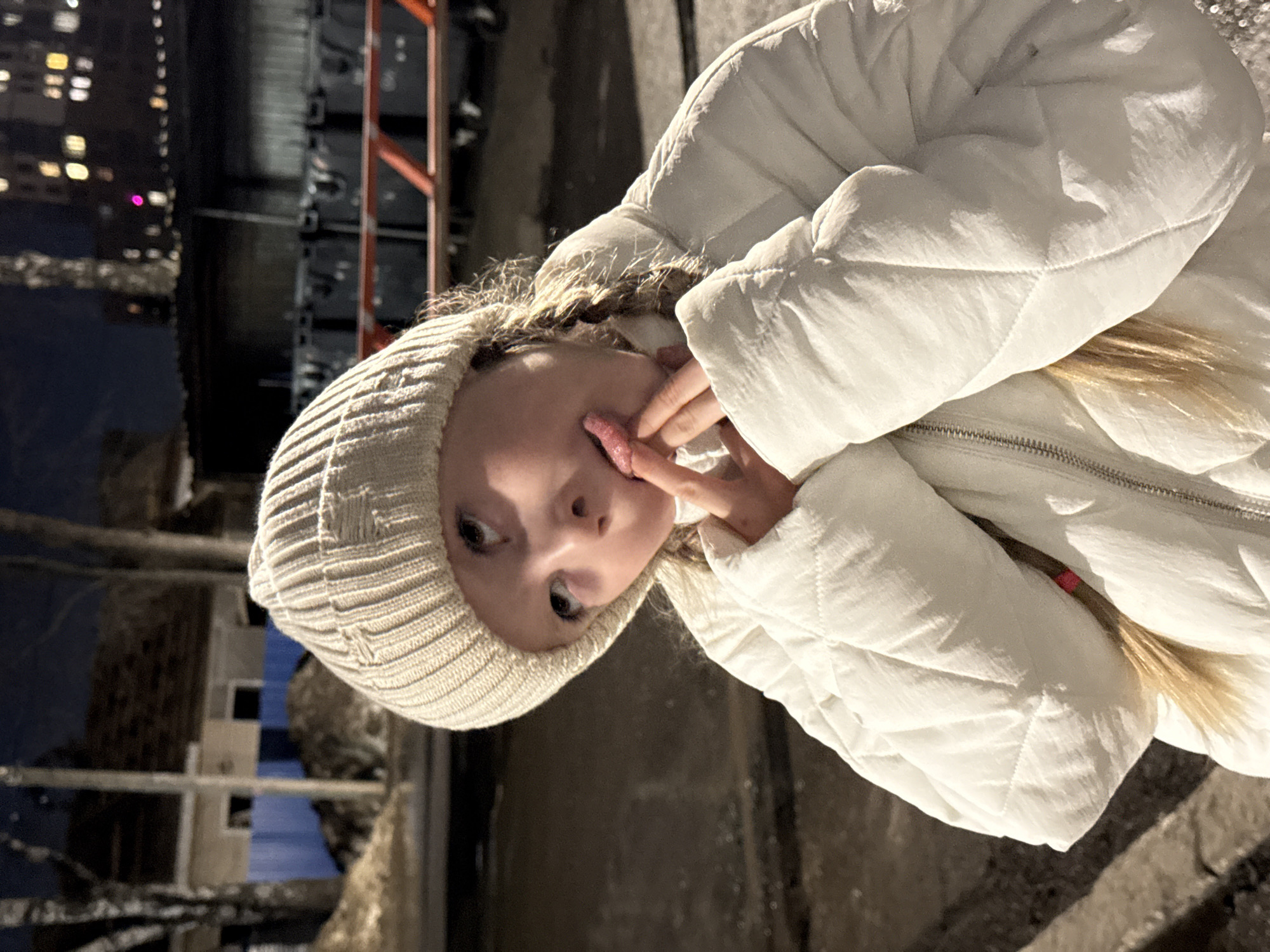 A child in a white winter coat and knitted cap is making a funny face, sticking out their tongue and pulling at their cheek with fingers. The background shows a city environment at night, with buildings visible in the distance. Details suggest a location in Izhevsk, Russia, with identifiable urban features. There is a red metal structure in the background, possibly scaffolding or a fence. A tree is partially visible in the foreground.