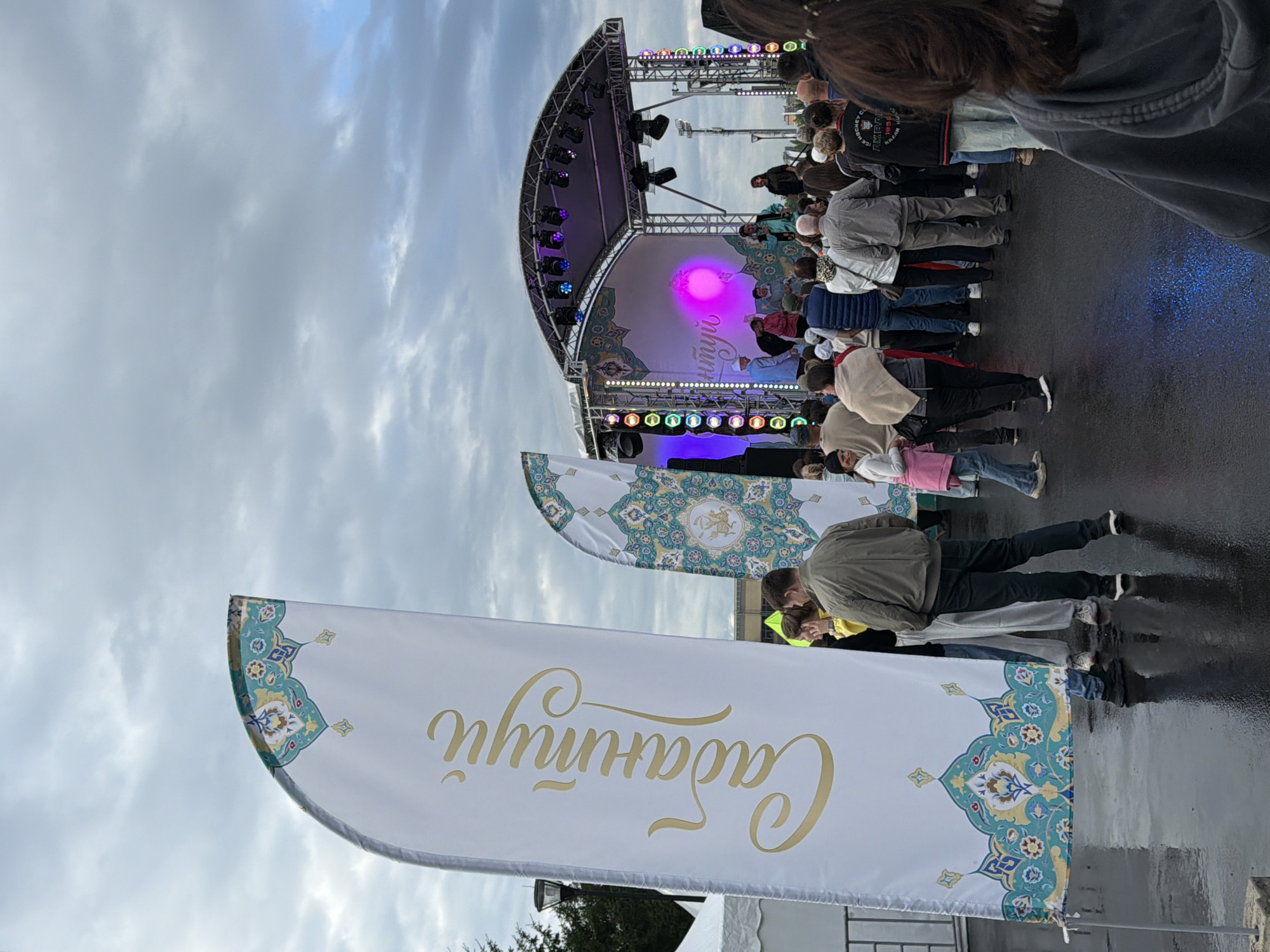 A group of people are gathered in an outdoor urban setting, possibly at a festival or event. In the foreground, a large white banner with gold calligraphy and intricate teal and yellow Islamic-inspired patterns is prominently displayed on the left. To the right of this banner, a smaller, similar banner is visible. Behind these banners, a crowd of people stands on a wet, reflective surface, looking towards a stage setup. The stage is illuminated with colorful lights, including purple and pink hues, and features a band or performers. The sky above is overcast with dramatic clouds. The overall atmosphere suggests an outdoor cultural or musical event.