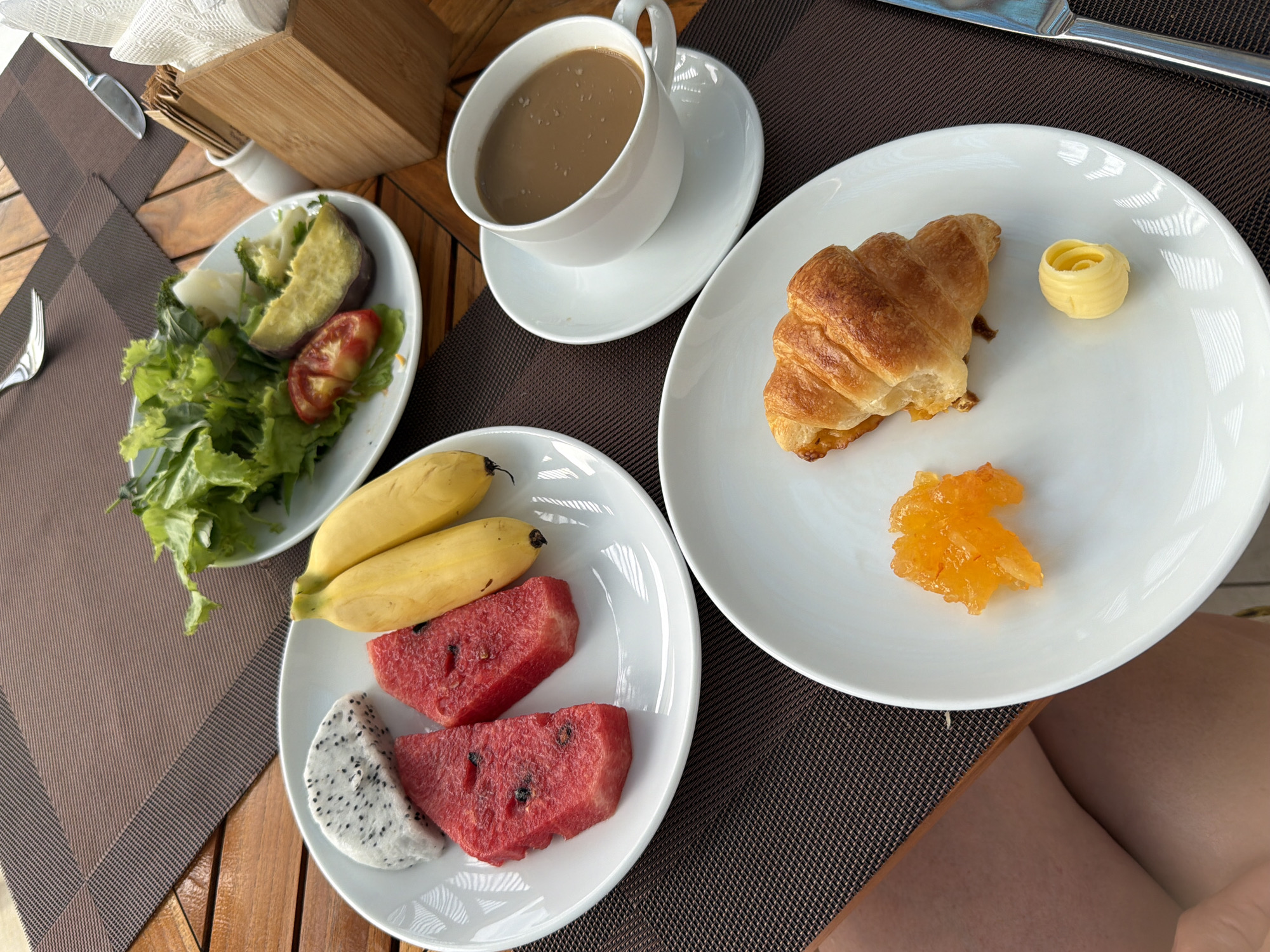 This is a top-down, medium shot of a breakfast setting on a wooden table with a brown placemat. In the foreground, a white plate contains a croissant, a swirl of butter, and a dollop of orange marmalade. To the left, another white plate holds slices of watermelon, two small bananas, and a slice of dragon fruit. Behind these plates, a larger white plate displays a salad with lettuce and tomato, accompanied by a piece of steamed sweet potato and broccoli. A white cup and saucer filled with coffee are positioned to the right of the fruit plates. Cutlery, a napkin holder with napkins, and a salt shaker are also visible on the table. The scene is set outdoors, likely at a restaurant in Dong Ninh Hoa Ward, Vietnam. The lighting suggests it is daytime. No people are visible in the frame.