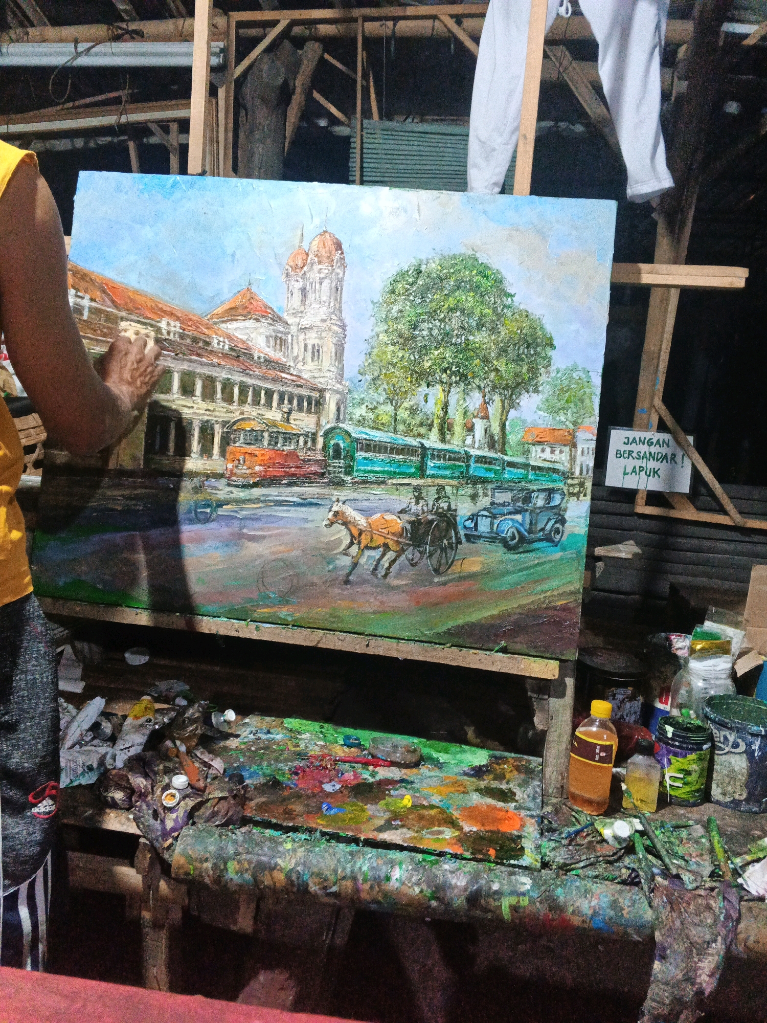 A person wearing a yellow shirt and patterned pants is in the process of painting an oil painting on an easel. The painting depicts a street scene with a building that has a dome-shaped tower, a large tree, several streetcars, a horse-drawn carriage, and an old-fashioned car. The setting appears to be outdoors, possibly in a workshop or studio, with wooden structures visible in the background and a cluttered workbench filled with art supplies in the foreground. A sign with Indonesian text, "JANGAN BERSANDAR! LAPUK!", is attached to a wooden beam to the right of the painting. The environment suggests an artistic endeavor in progress. The location is identified as Ungaran, Indonesia.