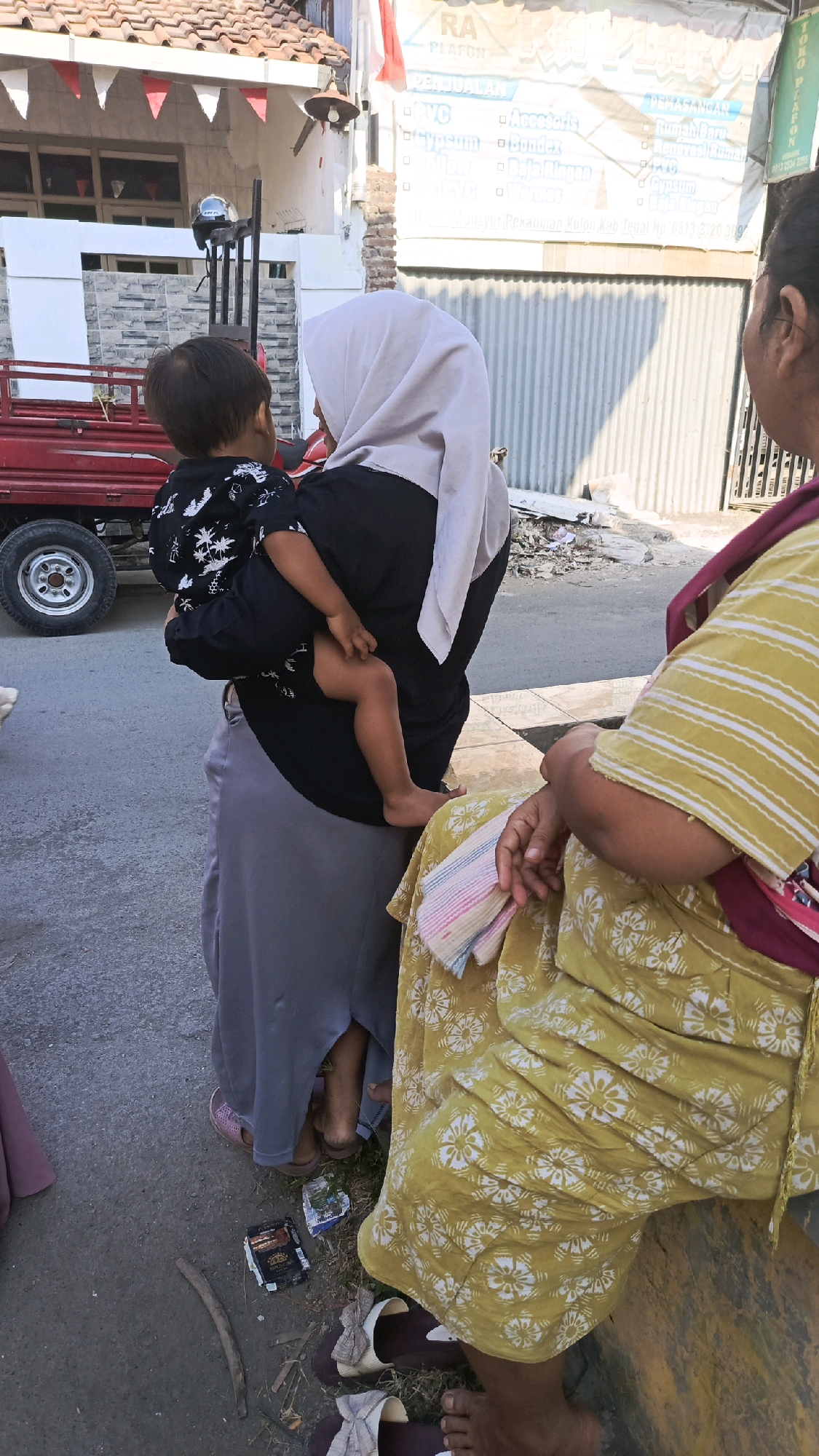 In Pekauman Kulon, Indonesia, a woman wearing a light grey hijab and a black top is holding a young child on her hip. The child is wearing a black shirt with a white palm tree print. The woman is also wearing a long grey skirt. To the right of the woman, another person is seated, wearing a yellow and white striped shirt and a patterned yellow skirt. The background shows a building with a red-tiled roof and white trim. A red vehicle is visible to the left of the frame, with a black tire. The ground is paved and there is some debris scattered around. A sign with Indonesian text is visible on the building. The lighting suggests it is daytime.