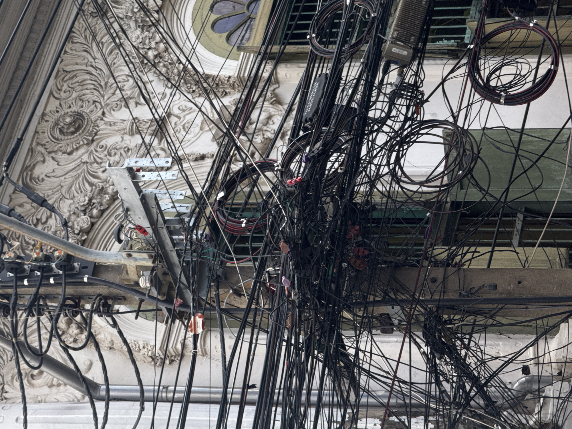 The image depicts a complex and dense network of electrical and telecommunication wires, a common sight in some urban areas. These wires are heavily tangled and wrapped around a utility pole. In the background, a building with ornate architectural details, possibly plaster or stone carvings, is visible. The building features an arched window with stained glass elements. The scene is likely from Bangkok, Thailand, where such intricate wire arrangements are characteristic of the city's infrastructure. No people are visible in the image.