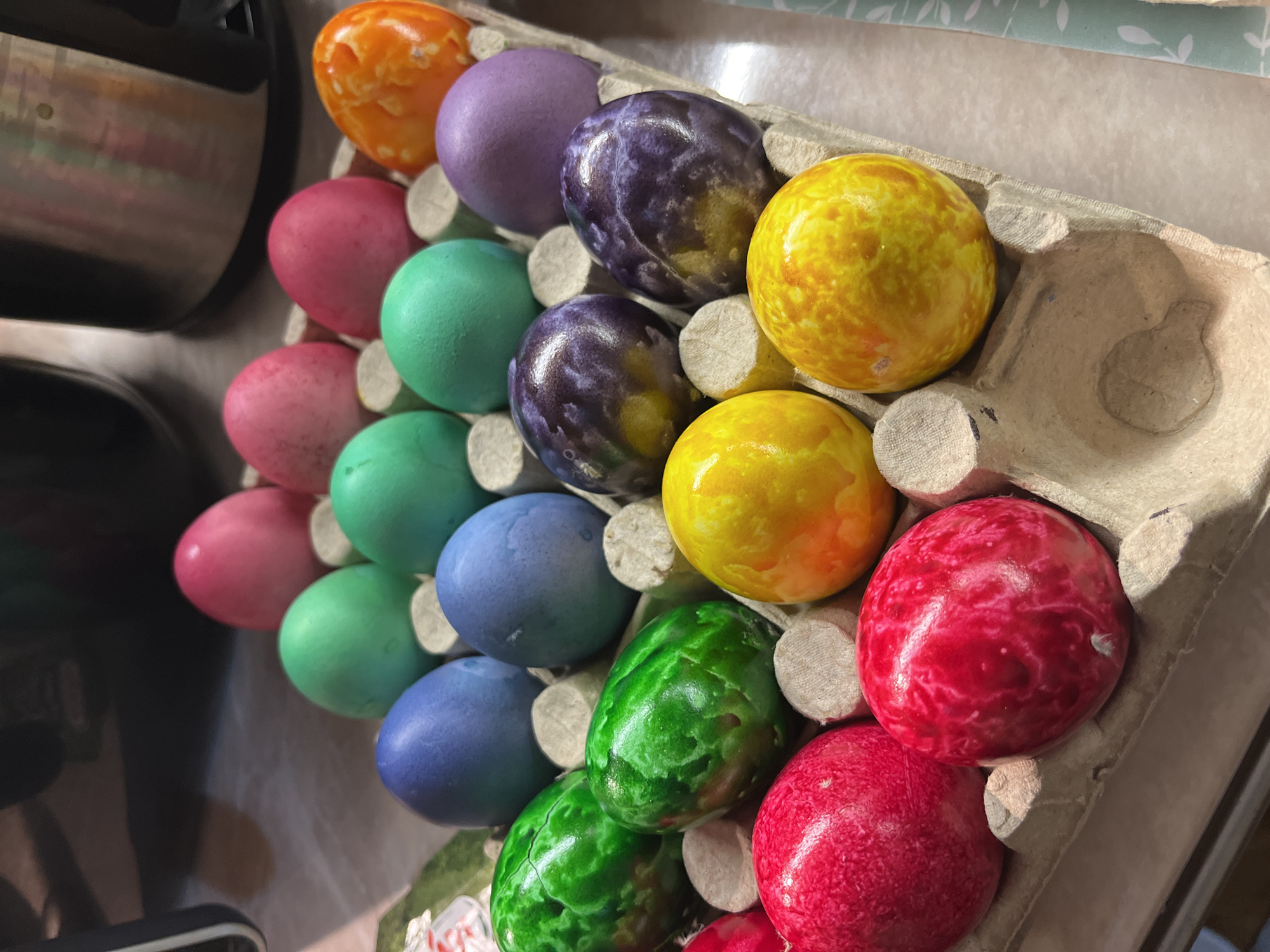 A close-up shot shows a cardboard egg carton filled with 24 dyed Easter eggs. The colors include orange, purple, dark purple, yellow, pink, light blue, green, and red. The eggs have a marbled appearance. The carton is partially filled, with some slots empty. The location and city are unknown.