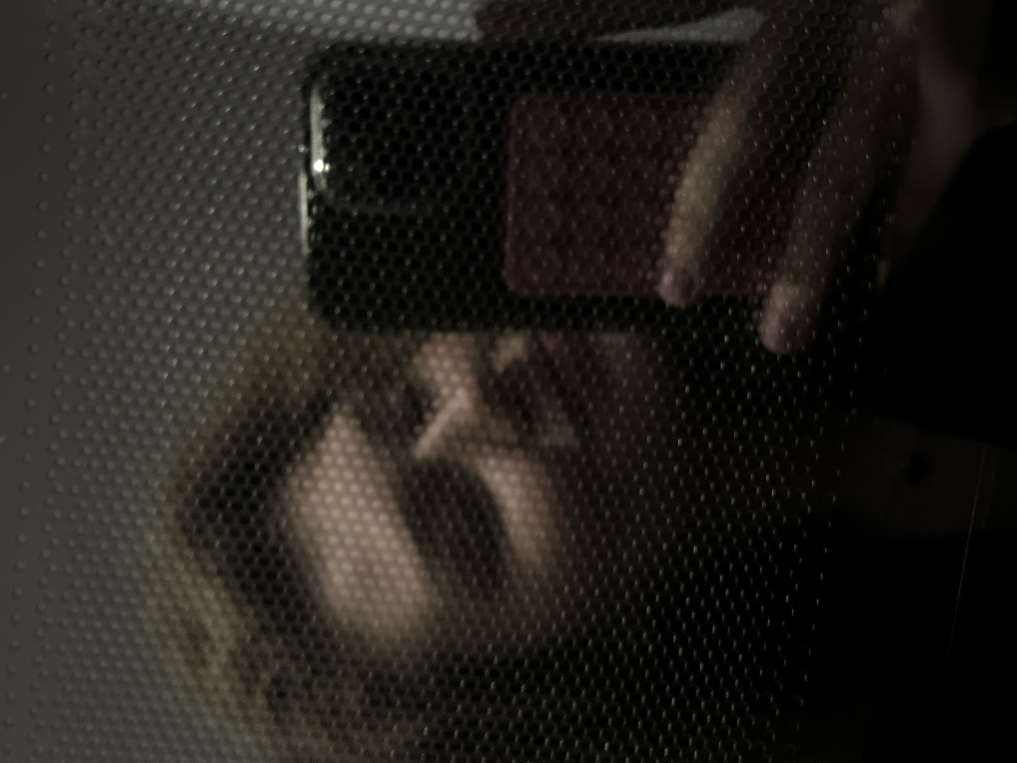 The image shows a person taking a selfie with a cell phone through a perforated surface. The person's face is partially visible in the reflection, obscured by the texture of the perforated surface. The cell phone is held in the upper center of the frame, with fingers visible around the device. The background is indistinct and dark. There is no information in the image to determine that the location is Kazan, Russia.