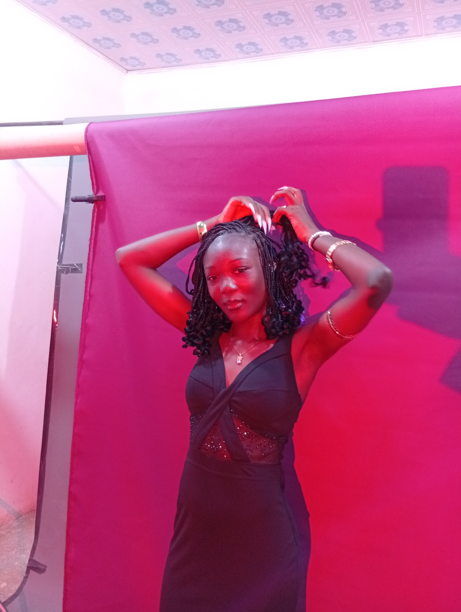 A young Black woman with braided hair is posing for a photo in front of a pink backdrop. She is wearing a black dress with a V-neckline and a sheer inset with beading on the bodice. She has on jewelry including bracelets on her right arm and a necklace. Her arms are raised, and she is touching her hair. The lighting is predominantly pink, casting shadows on the backdrop. The ceiling above the backdrop appears to be a dropped ceiling with decorative tiles. The location is an indoor setting.