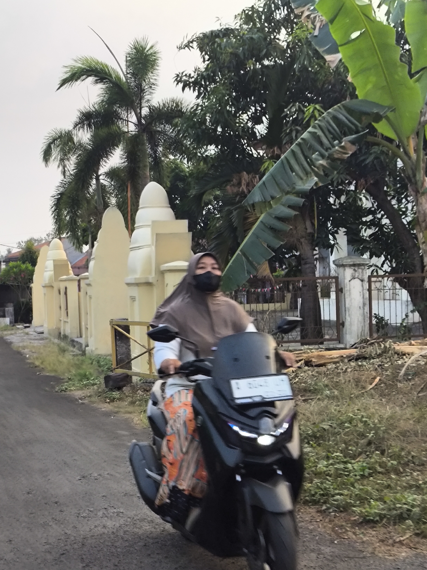 A woman wearing a brown hijab and black face mask is riding a black motorcycle on a dirt road. She is wearing a white long-sleeved shirt and a colorful fabric with orange, brown, and white patterns is draped over her legs. The motorcycle has a white license plate with blue and white lettering. The background features a yellow wall with decorative white tops, palm trees, and lush green vegetation. The scene appears to be in a rural or suburban area of Cilegon, Indonesia, possibly near a residential area or a place of worship given the architectural style of the wall. The lighting suggests it is late afternoon or early evening.