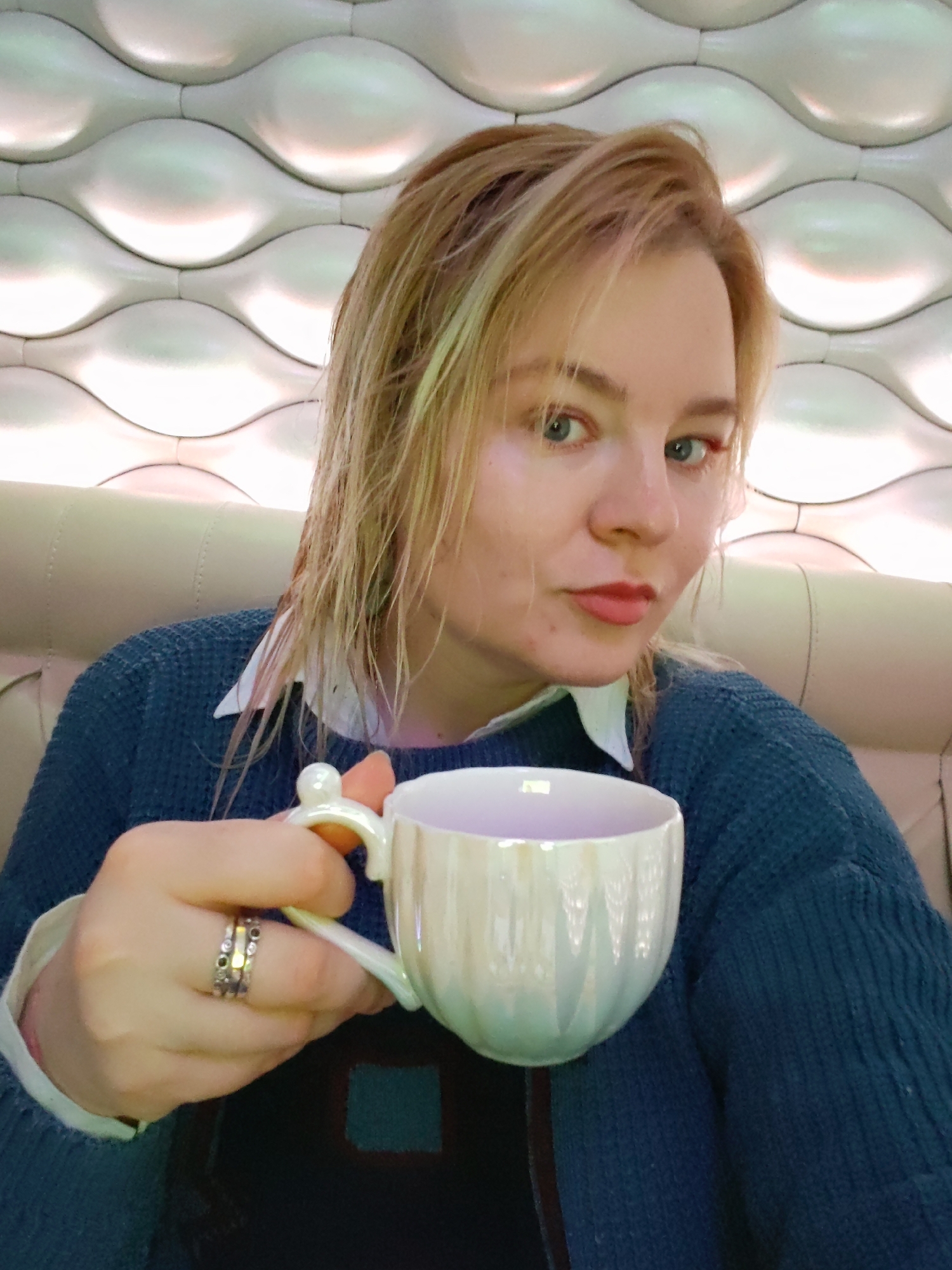 A fair-skinned woman with wet, blonde hair is seated inside what appears to be a cafe or restaurant. She is holding a pearlescent white tea cup with a decorative handle. She wears a blue sweater over a white collared shirt, and rings are visible on her hand. The background shows a wall with a repeating, wave-like pattern and a beige seat. The photograph's composition focuses primarily on the woman and the cup she is holding. It is unknown if this photo was taken in Izhevsk, Russia.
