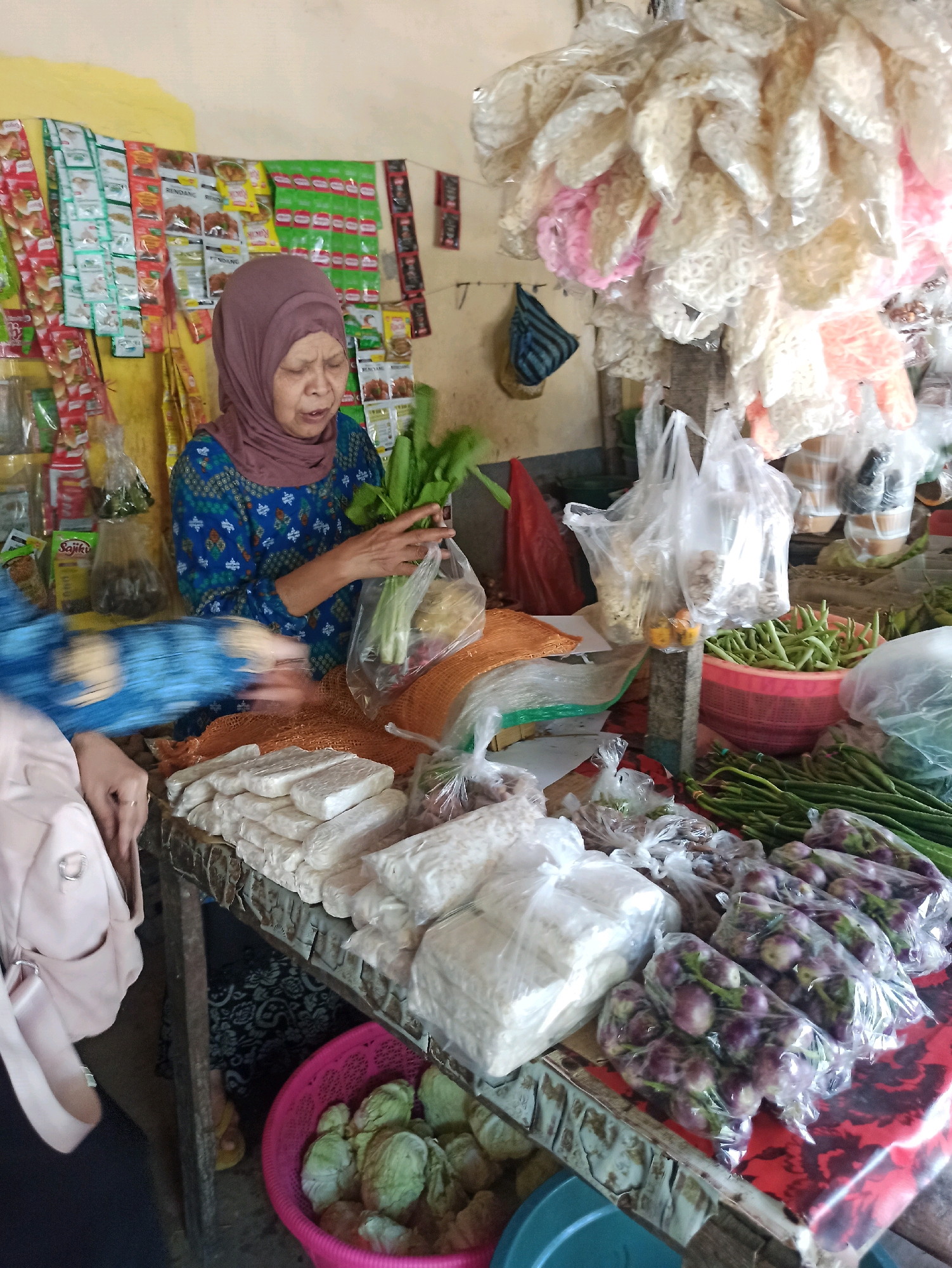 A middle-aged Indonesian woman wearing a purple headscarf and a blue patterned top is seen at a market stall in Tlogosari, Indonesia. She is holding a bunch of leafy green vegetables in plastic. In front of her, on a wooden counter, are stacks of white, rectangular items that appear to be tempeh, along with other packaged food items and produce. To the right, there are baskets of green beans and bundles of long green beans, as well as plastic-wrapped eggplants. The background shows shelves stocked with packaged spices and snacks. The overall scene depicts a vibrant market environment with a focus on fresh food and groceries.