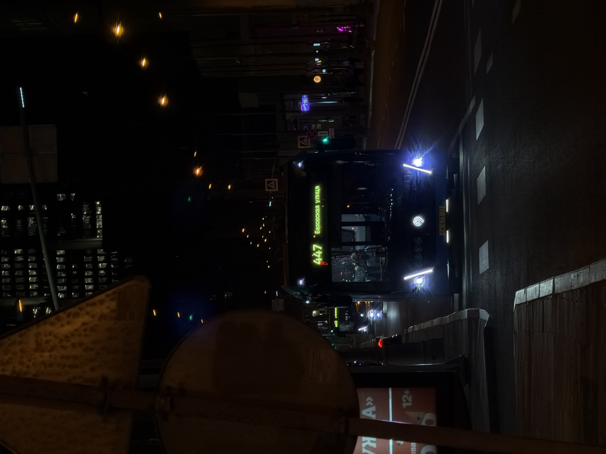 A nighttime street scene in Moscow, Russia, features a bus with the number 447 displayed, its destination sign reading "Базовская улица" (Bazovskaya Street) in green lettering. The bus' headlights are illuminated. The street is dark with reflections from lights on the wet pavement. Additional streetlights line the road. A sign or advertisement with the text "A" and "12+" is visible in the foreground. In the background, other city lights and building windows suggest an urban setting. A metal object, possibly part of a street sign or structure, is prominently displayed in the lower left corner of the image.