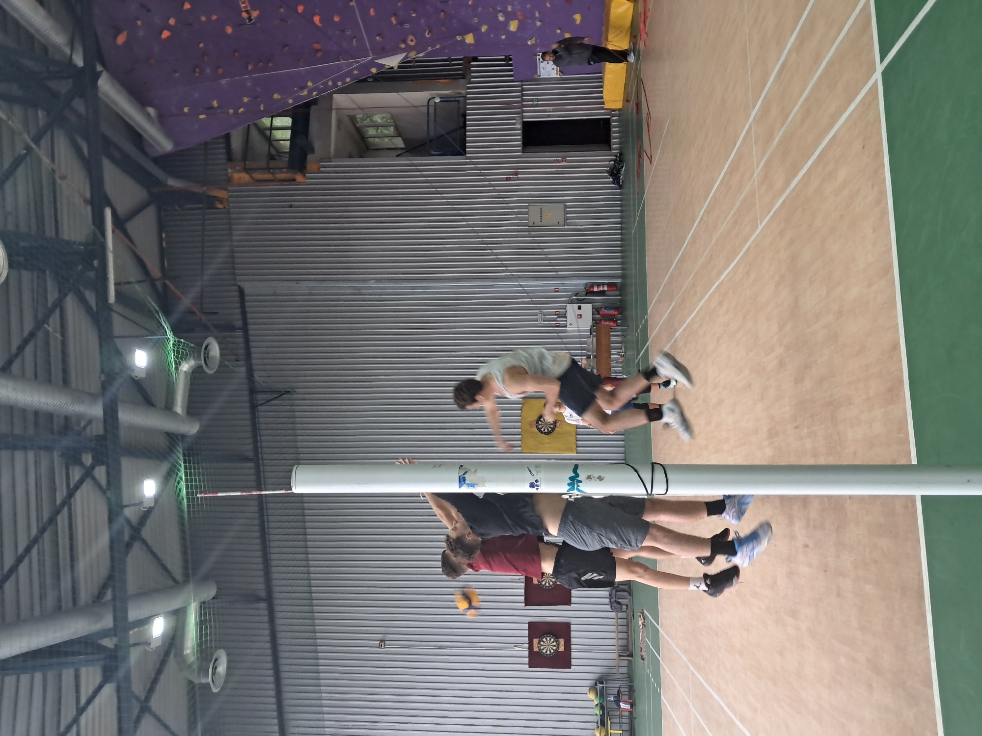 This image captures a dynamic scene inside a multi-purpose indoor sports facility, likely located in Veliko Tarnovo, Bulgaria, as indicated by the provided context. The perspective is from a slightly elevated position, looking across a sports court.
The primary subjects are three male individuals engaged in a game of volleyball or a similar net sport. Two players are positioned centrally near a tall white net pole, both in mid-air, indicating they are jumping or spiking. One wears a red t-shirt and black shorts, the other a black t-shirt and grey shorts. A yellow and blue volleyball is suspended in the air near them, suggesting active play. A third player, in a light-colored t-shirt and dark shorts, is further back on the court, also appearing to be in motion. In the far background, another individual in dark clothing stands on a platform next to a purple climbing wall.
The setting is an indoor sports hall with a light brown court marked with white lines, bordered by a green area. The walls are constructed from corrugated metal panels, and the ceiling features exposed structural beams, ventilation ducts, and bright overhead lights. In the background, a large purple climbing wall with numerous orange holds is visible, suggesting the facility caters to various sports. Notable objects include multiple dartboards mounted on the corrugated wall, and various pieces of sports equipment like balls and bags near the perimeter. On the white net pole, the text "CTPOH" and some stylized graphic elements are discernible. The lighting suggests an indoor daytime scene, relying on artificial illumination. The overall atmosphere is one of active sports engagement within a modern, functional facility.