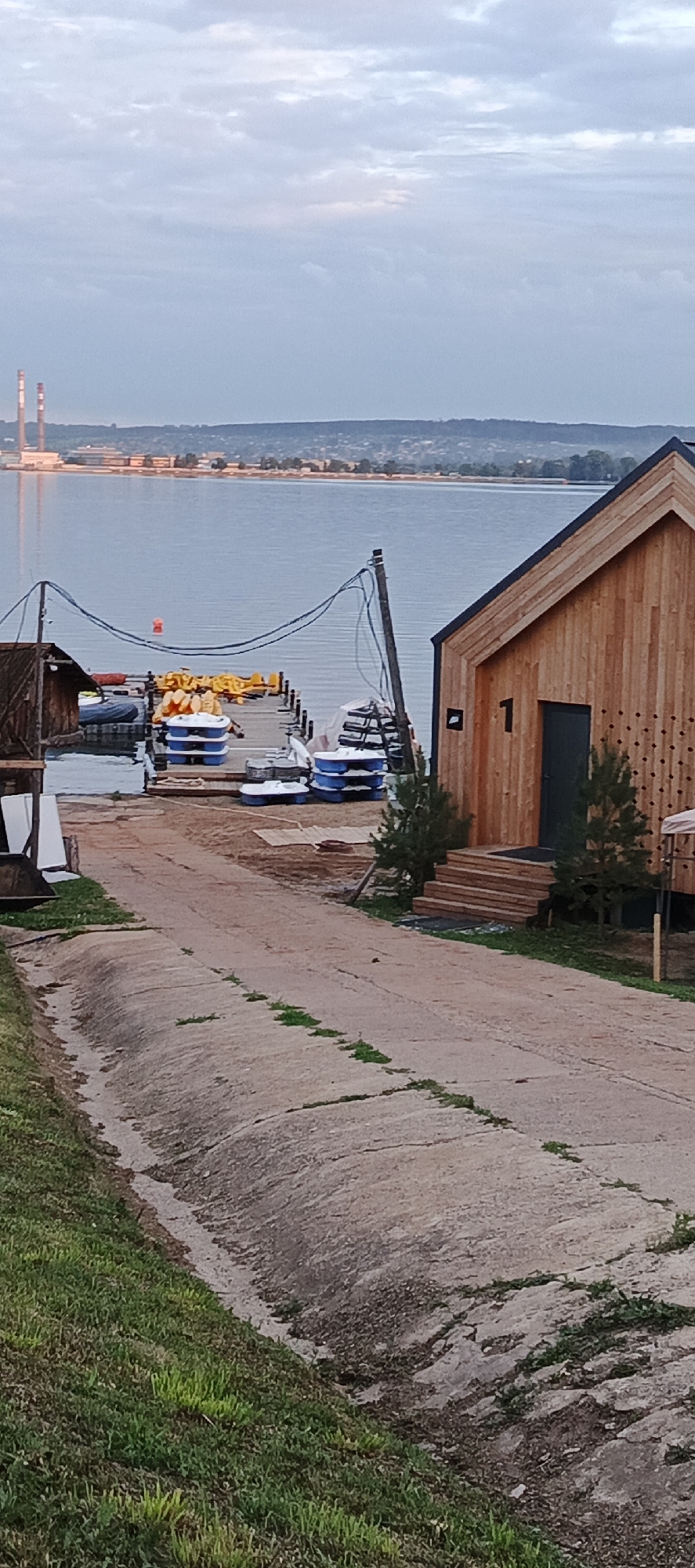 The image depicts a waterfront scene in Votkinsk, Russia, likely at a recreational area or marina. In the foreground, a sloped, cracked concrete ramp leads down to a wooden dock. The dock is lined with several blue and white pedal boats and some yellow floatation devices. To the right of the ramp, a modern wooden building with a dark door and a prominently displayed number "1" is visible, suggesting it might be accommodation or a rental facility. A small set of wooden steps leads up to its entrance, flanked by two coniferous trees.
The middle ground features a calm body of water, a lake or river, with the Votkinsk cityscape and industrial structures, including two large chimneys, visible in the distance across the water. The sky is overcast with soft, diffused light, indicating it is either early morning or late evening. The overall impression is one of a tranquil, possibly undeveloped, waterfront area. There are no people visible in the image.