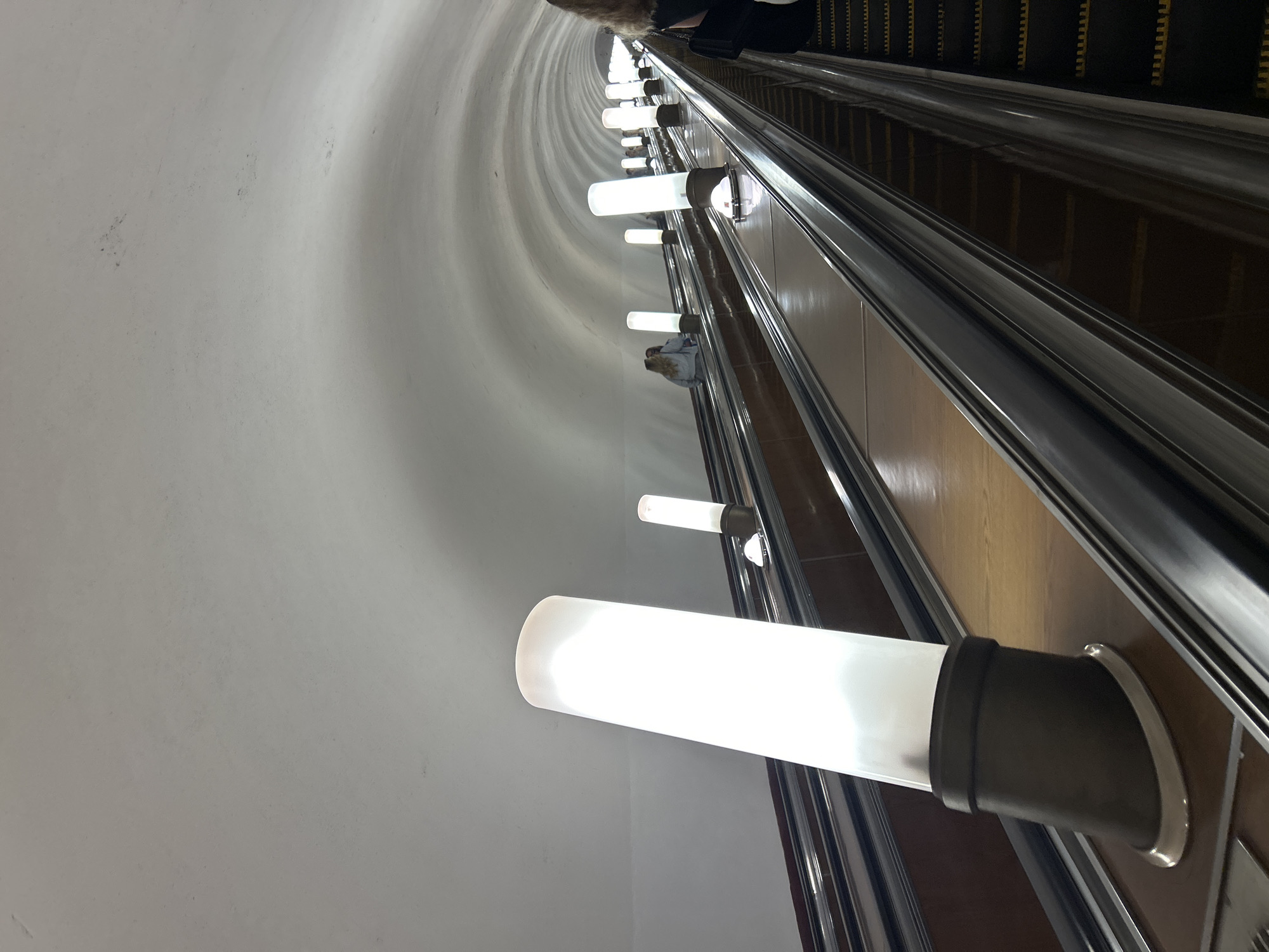 An upward-angled, wide shot shows a long, curved escalator tunnel with parallel escalators. The tunnel ceiling is a smooth, off-white curve, illuminated by evenly spaced, tall, cylindrical white lights on the sides of the escalators. The escalators themselves have dark grey metal handrails and treads, with wooden-effect sides. Several people are visible on the escalators, ascending. In the foreground, a large, bright white cylindrical light fixture is prominent on the left escalator. Further down the tunnel, more people and lights are visible, receding into the distance and the tunnel's curve. The perspective creates a sense of depth and the vastness of the underground space.