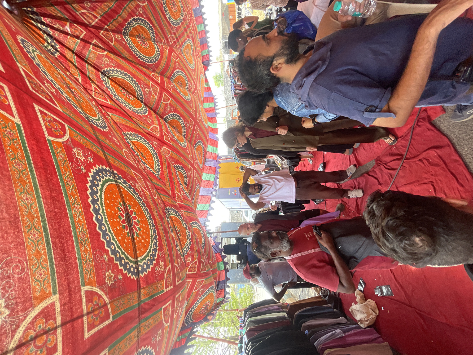 The image shows a gathering of people under a large, red, patterned canopy, likely in a market or public space. The location is Anajpur, India.
People:
- A group of men of varying ages are visible.
- They are standing and sitting on a red fabric or covering.
- Some appear to be looking at something or interacting with each other.
- Some are wearing casual clothing such as t-shirts, pants, and jeans.
Objects:
- A large, red canopy with a circular patterned design covers the area.
- Water bottles are visible.
- A collection of clothing items is stacked near the bottom of the frame.
- A small number of electronic devices are in use
Actions:
- People are standing and sitting, some talking to each other
- Some appear to be taking a break or resting.
Environment:
- The scene appears to be an outdoor location, possibly a market or a temporary setup.
- There are glimpses of buildings and a vehicle in the background.
- The overall impression is that the gathering is part of a market, event, or protest.