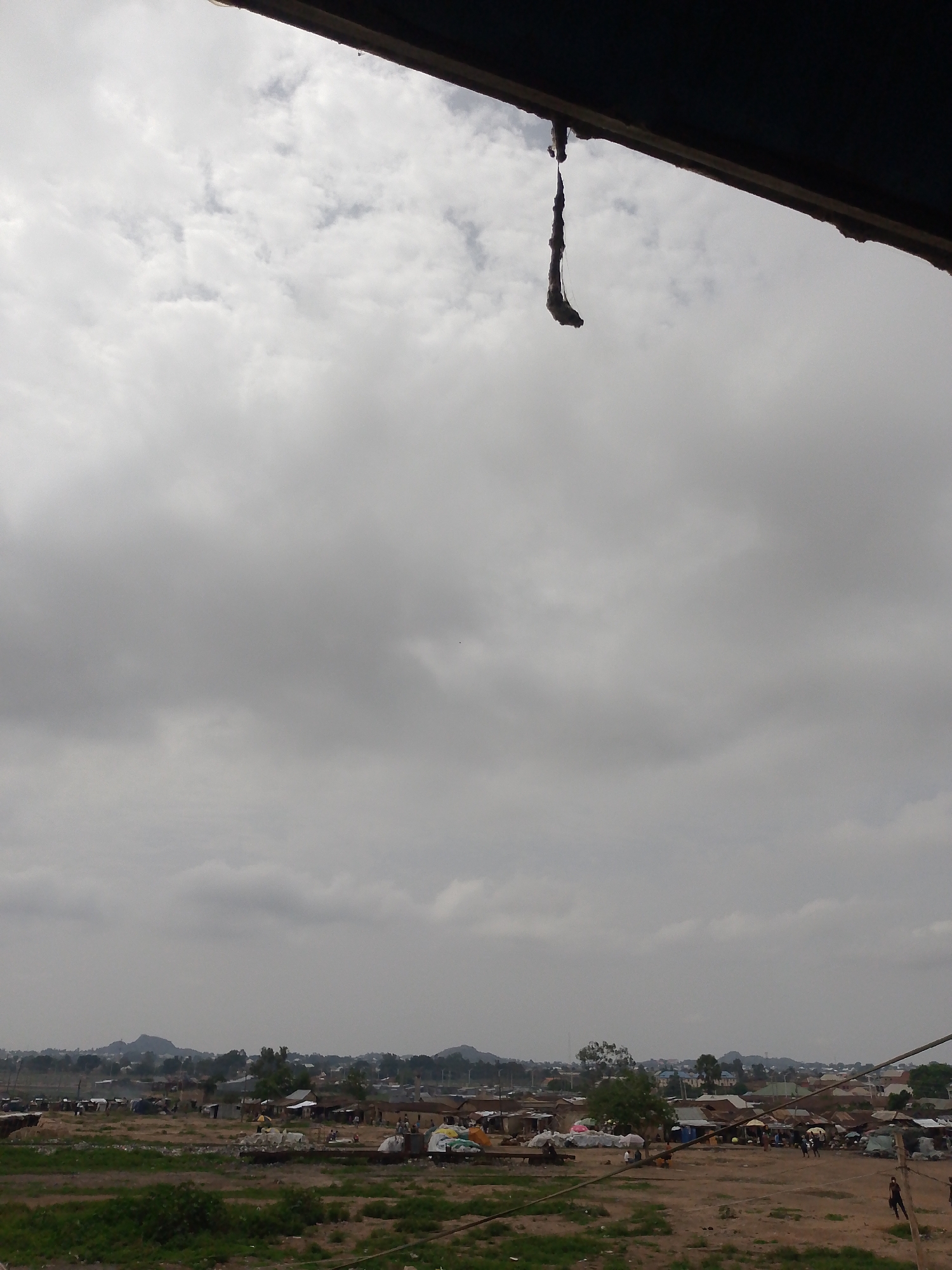 The image depicts a view from under a roof structure overlooking a residential area in Bukuru, Nigeria. The sky is overcast. In the distance, there are rolling hills. The residential area appears to have modest housing, with a mix of structures and open spaces. There are people present in the scene. In the foreground, the ground is mostly dirt and sparse vegetation. A hanging debris is visible from the roof structure.