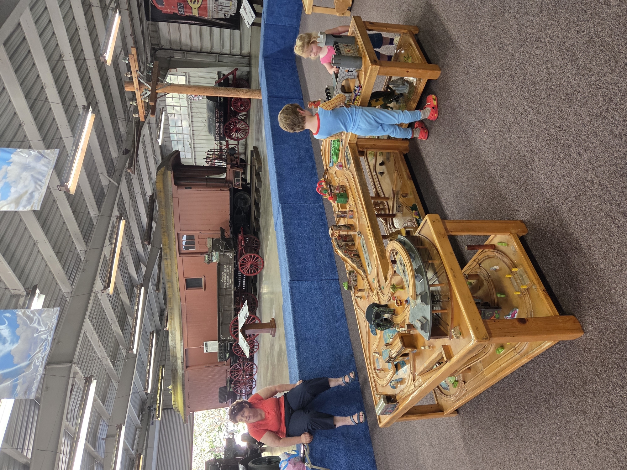 A woman wearing a coral t-shirt and dark pants sits on a blue cushioned bench inside a large building, possibly a museum or exhibition hall. She is looking to her left with a pensive expression. In the foreground, two young children, a boy in a blue jumpsuit and a girl in a pink shirt, are playing with a large wooden train set. The train set is elaborate, with tracks, bridges, tunnels, and various miniature buildings and accessories. The building's interior features a high ceiling with exposed beams and fluorescent lighting. In the background, a vintage red train car with large red wheels is parked on tracks, and other historical railway equipment is visible. The floor is covered with dark brown carpet. The overall impression is of a family-friendly attraction, likely related to trains or transportation history. The location is not specified in the image, so it cannot be determined to be in Los Angeles, United States of America.