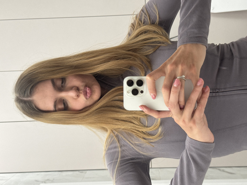 A woman with long blonde hair takes a selfie in a mirror. She wears a grey long-sleeved shirt. Her lips are puckered in a kiss. She holds a white smartphone with three visible camera lenses. A decorative ring adorns one of her fingers. The background features a white wall and a glimpse of marble flooring.