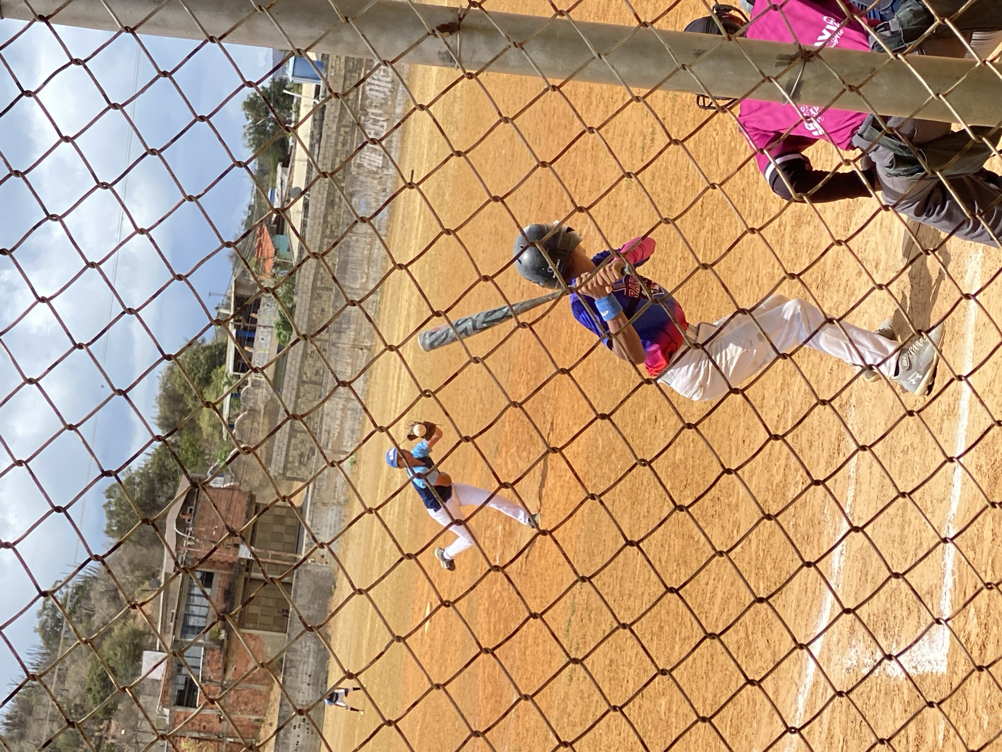 The image captures a baseball game in progress, viewed through a chain-link fence. A batter in a blue and pink uniform swings at a pitch, while a fielder in blue prepares to catch the ball. An umpire in pink stands nearby. The game takes place on a clay field with stadium seating in the background, amidst buildings and foliage. The location is Altagracia, Venezuela.