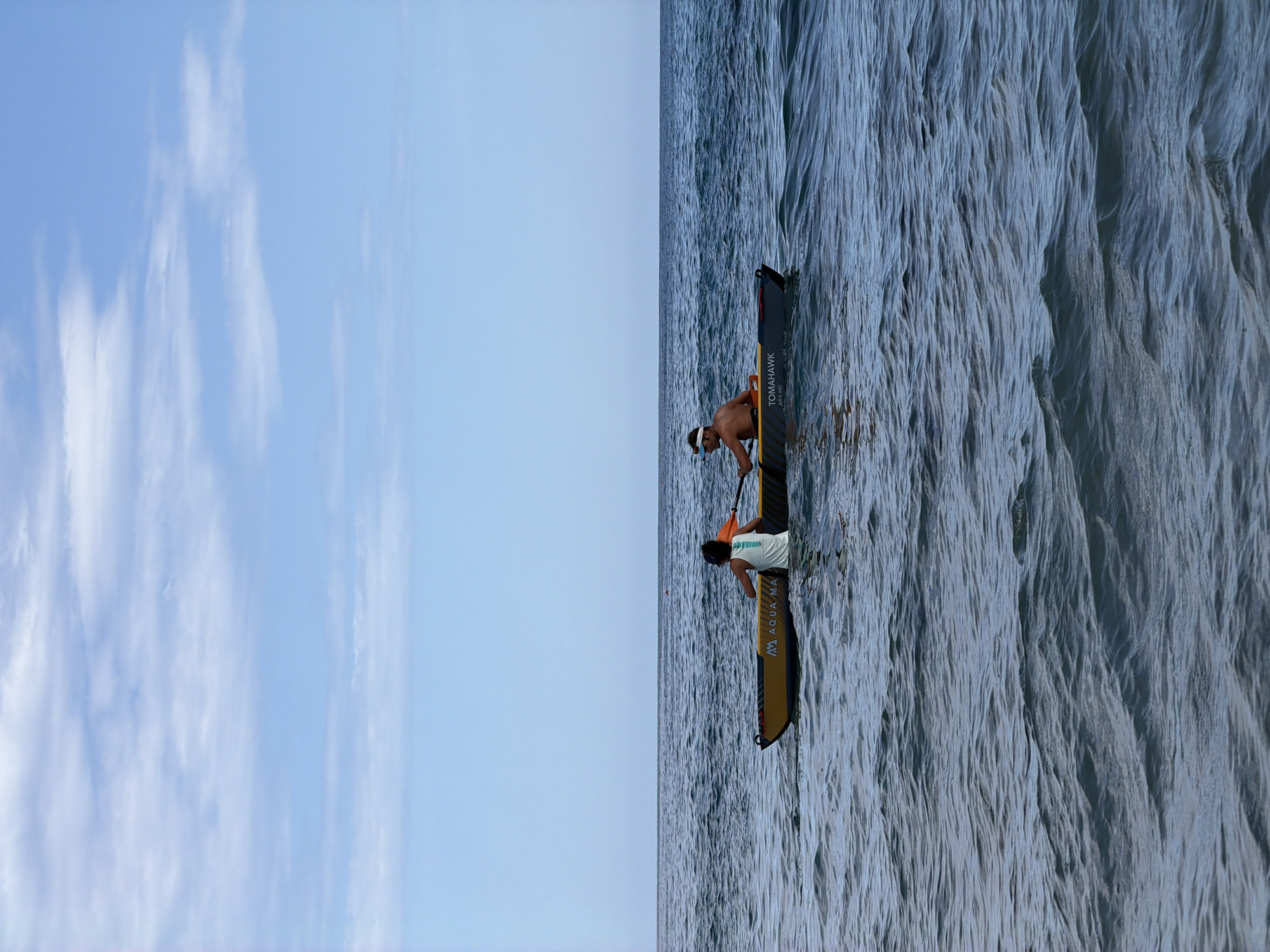 Two individuals are kayaking on the sea under a partially cloudy blue sky. The kayak is yellow and gray with "TOMAHOWK AIR-K 440" printed on the side, along with a logo that appears to be "Aqua-Ma". The person in the front is wearing a white tank top with an orange design and a dark headband. The person in the back is wearing a white visor and is shirtless. Both are holding paddles, with the front person's paddle extending to the right and the back person's paddle extending to the left. The water is choppy with small waves. This image does not provide any identifiable features to confirm it is in Tortolì, Italy.